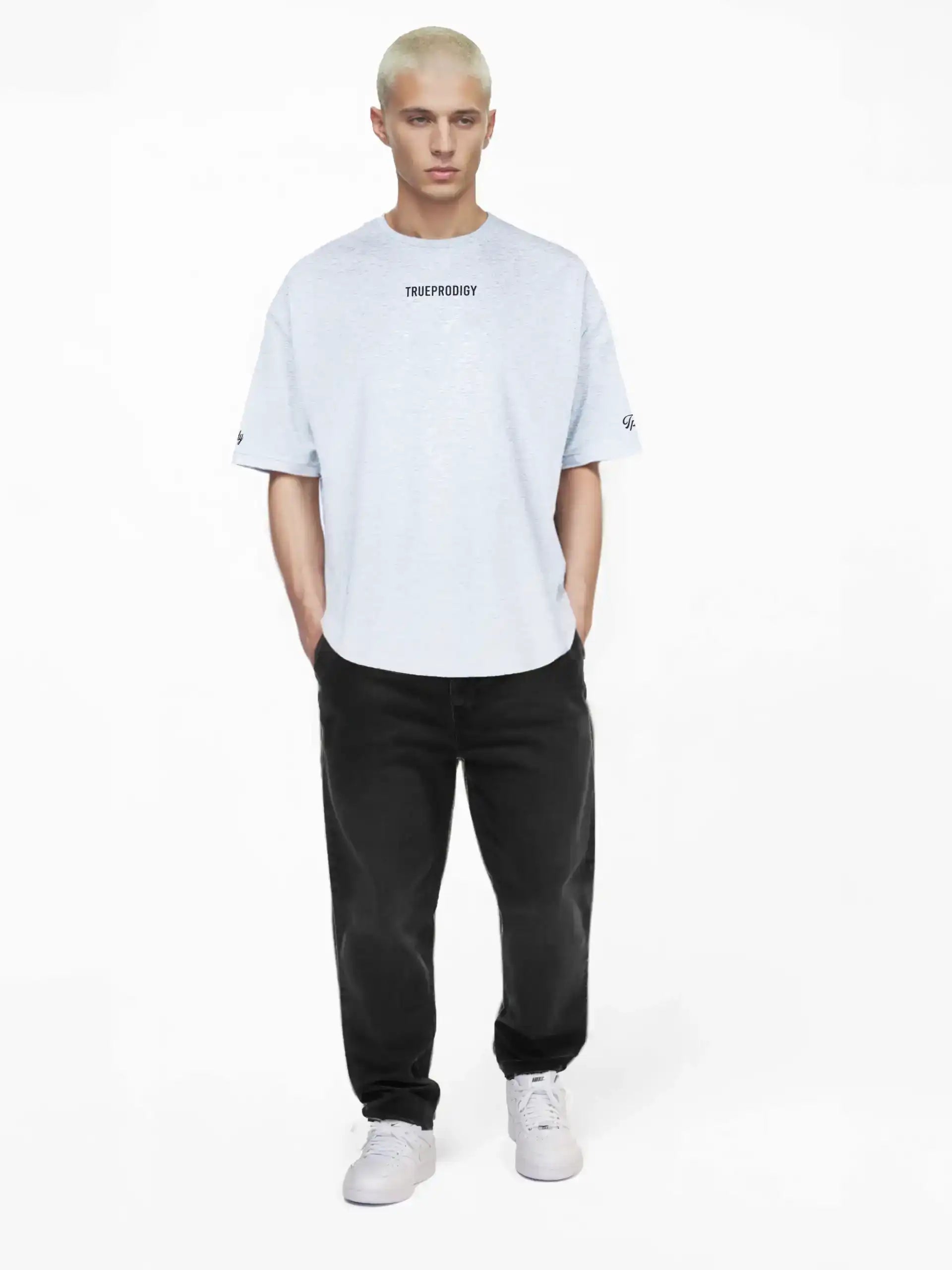Eine Person mit kurzen, gebleachten blonden Haaren steht vor einem weißen Hintergrund und trägt das trueprodigy Kai Oversize-Herren-T-Shirt mit "LIMITLESS"-Rückenaufdruck und Logo auf der Vorderseite, gepaart mit einer schwarzen Jeans und weißen Sneakers.