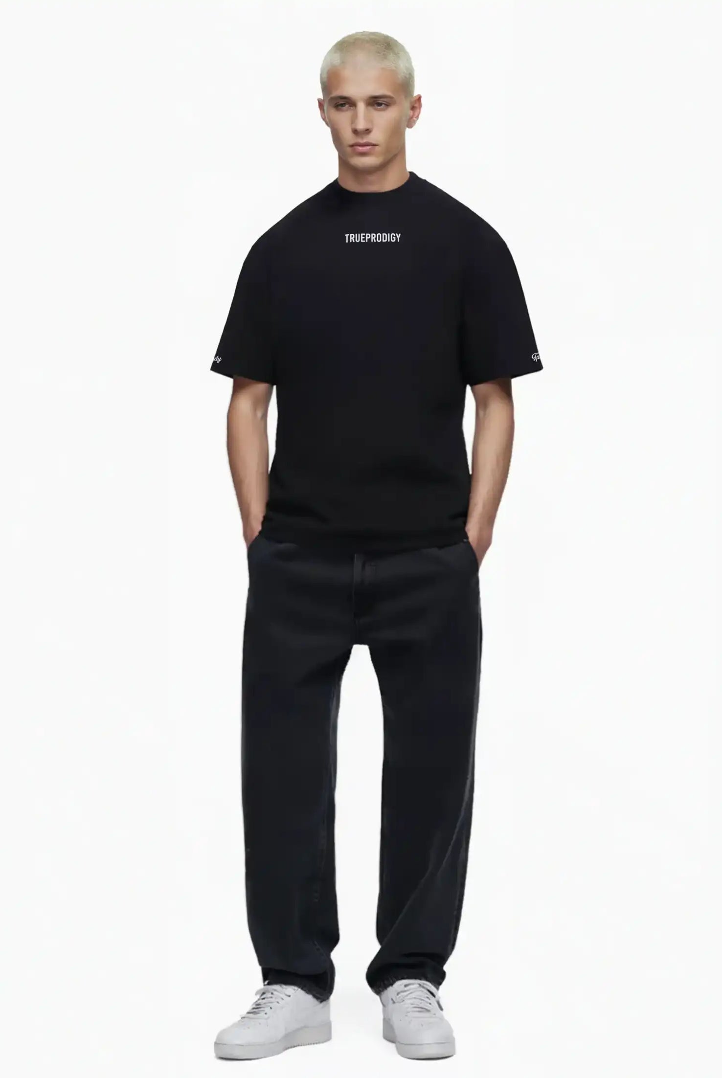 Eine Person mit kurzen blonden Haaren steht vor weißem Hintergrund und trägt das trueprodigy Kai Oversize-Herren-T-Shirt mit Rückenprint "LIMITLESS" und Markenlogo vorn, gepaart mit schwarzen Jeans und weißen Turnschuhen, die Hände in den Taschen.