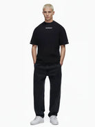 Eine Person mit kurzen blonden Haaren steht vor weißem Hintergrund und trägt das trueprodigy Kai Oversize-Herren-T-Shirt mit Rückenprint "LIMITLESS" und Markenlogo vorn, gepaart mit schwarzen Jeans und weißen Turnschuhen, die Hände in den Taschen.