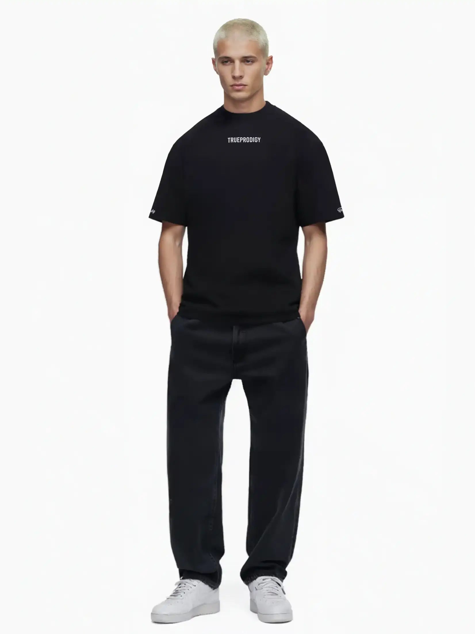 Eine Person mit kurzen blonden Haaren steht vor weißem Hintergrund und trägt das trueprodigy Kai Oversize-Herren-T-Shirt mit Rückenprint "LIMITLESS" und Markenlogo vorn, gepaart mit schwarzen Jeans und weißen Turnschuhen, die Hände in den Taschen.