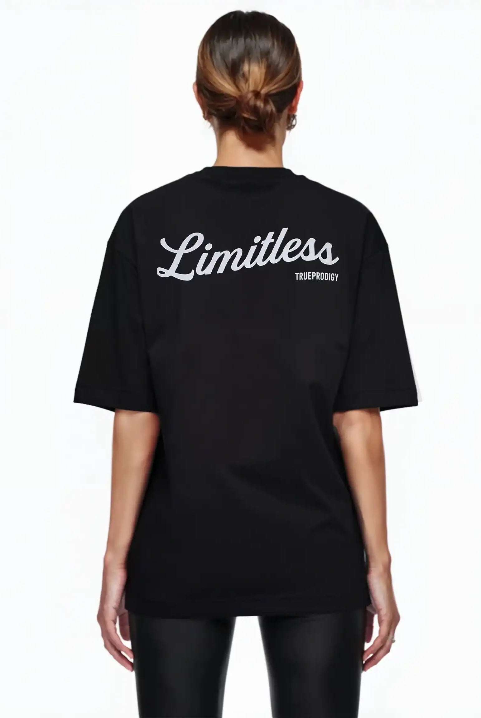 Eine Frau mit braunen Haaren in einem tiefen Dutt ist von hinten zu sehen. Sie trägt das Kai F Oversize-Damen-T-Shirt von trueprodigy mit einem "LIMITLESS"-Rückenaufdruck, gepaart mit einer schwarzen Hose vor einem schlichten weißen Hintergrund.