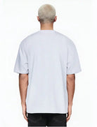 Gezeigt wird eine Person mit kurzen, gebleichten blonden Haaren von hinten, die ein hellgraues Ravi Oversize-Shirt mit großem trueprodigy-Schriftzug vorn by trueprodigy und eine schwarze Hose vor einem schlichten weißen Hintergrund trägt.