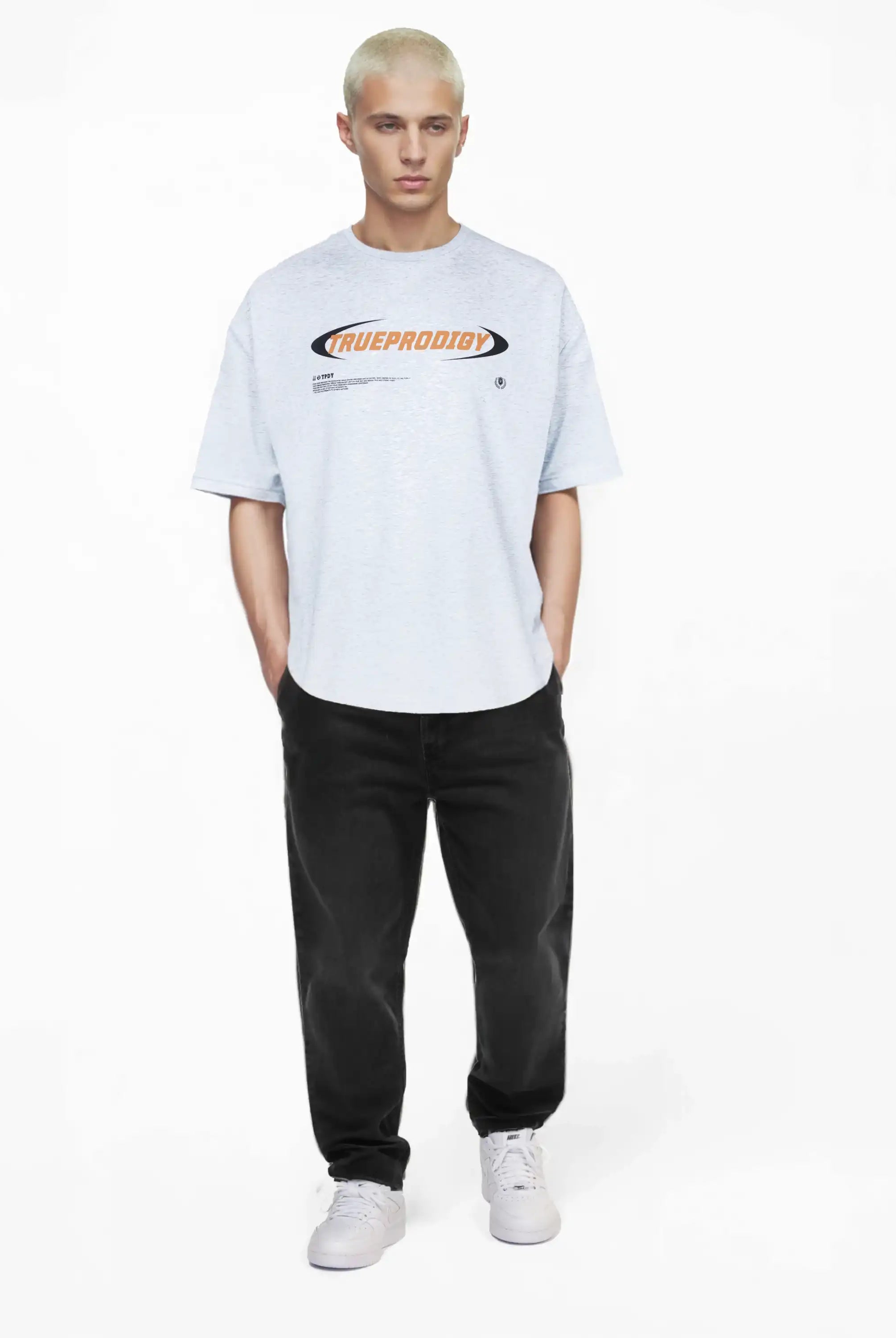 Eine Person mit kurzen, gebleichten blonden Haaren blickt nach vorne und trägt das trueprodigy Ravi Oversize-Shirt mit großem trueprodigy-Schriftzug vorn in Hellgrau, gepaart mit schwarzen Jeans und weißen Turnschuhen vor einem schlichten weißen Hintergrund.