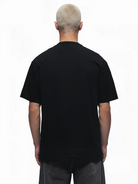 Eine Person mit kurzen, gebleichten Haaren steht mit dem Rücken zur Kamera, trägt das trueprodigy Ravi Oversize-Shirt mit großem trueprodigy-Schriftzug vorn und eine dunkelgraue schwere Jersey-Stoffhose vor einem schlichten weißen Hintergrund.