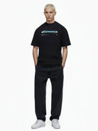 Eine Person mit kurzen blonden Haaren trägt das trueprodigy Ravi Oversize-Shirt mit großem Schriftzug, gepaart mit lockeren schwarzen Jeans und weißen Turnschuhen, die Hände in den Taschen vor einem schlichten weißen Hintergrund.