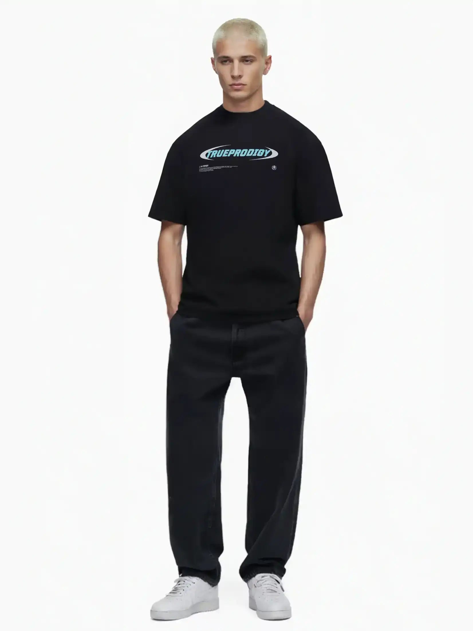 Eine Person mit kurzen blonden Haaren trägt das trueprodigy Ravi Oversize-Shirt mit großem Schriftzug, gepaart mit lockeren schwarzen Jeans und weißen Turnschuhen, die Hände in den Taschen vor einem schlichten weißen Hintergrund.