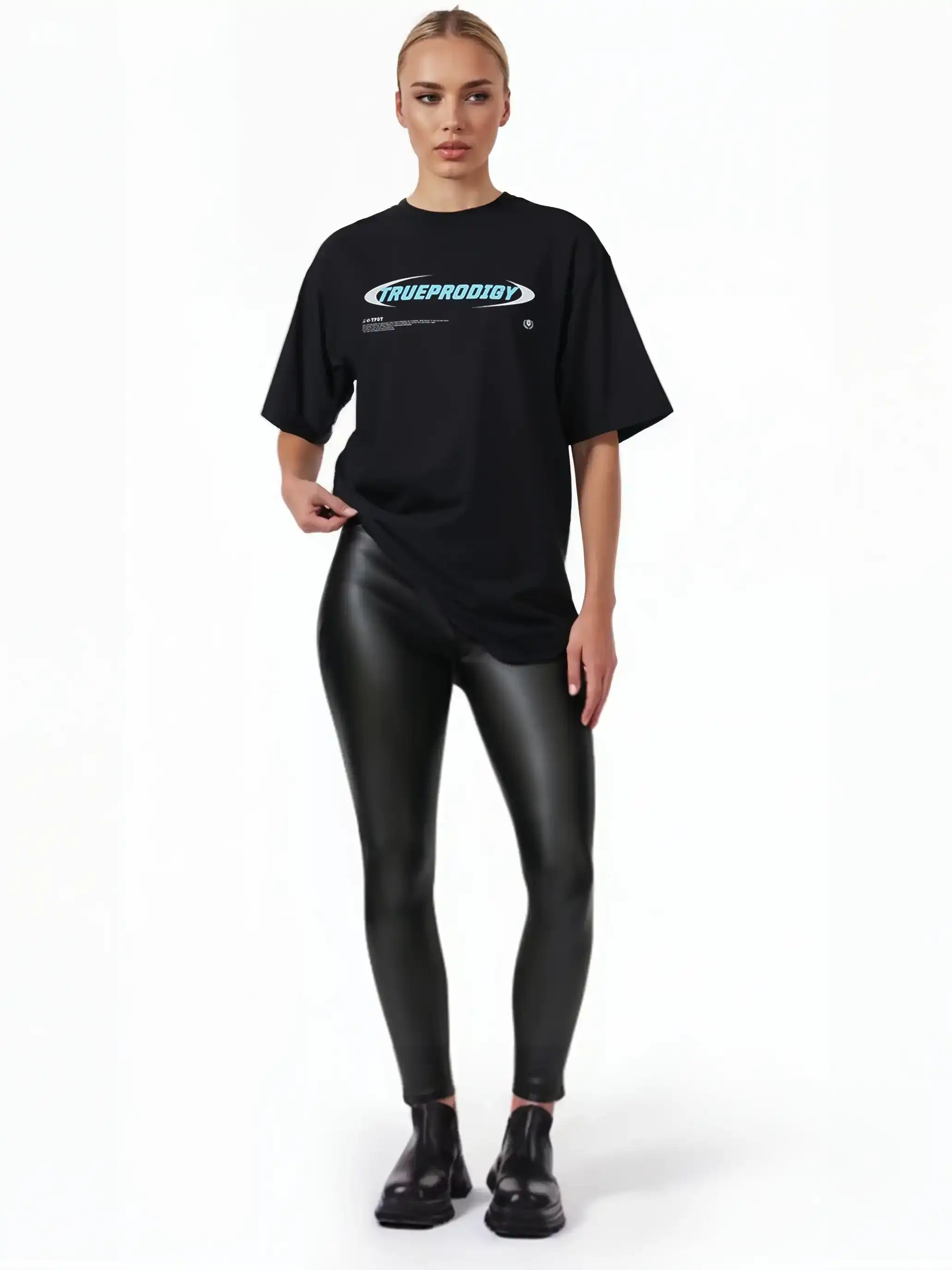 Eine Frau steht vor einem weißen Hintergrund und trägt das trueprodigy Ravi F Oversize-T-Shirt mit farbigem TRUEPRODIGY-Frontprint, schwarze Kunstlederleggings und schwarze Schuhe. Sie schaut mit neutralem Blick direkt in die Kamera.