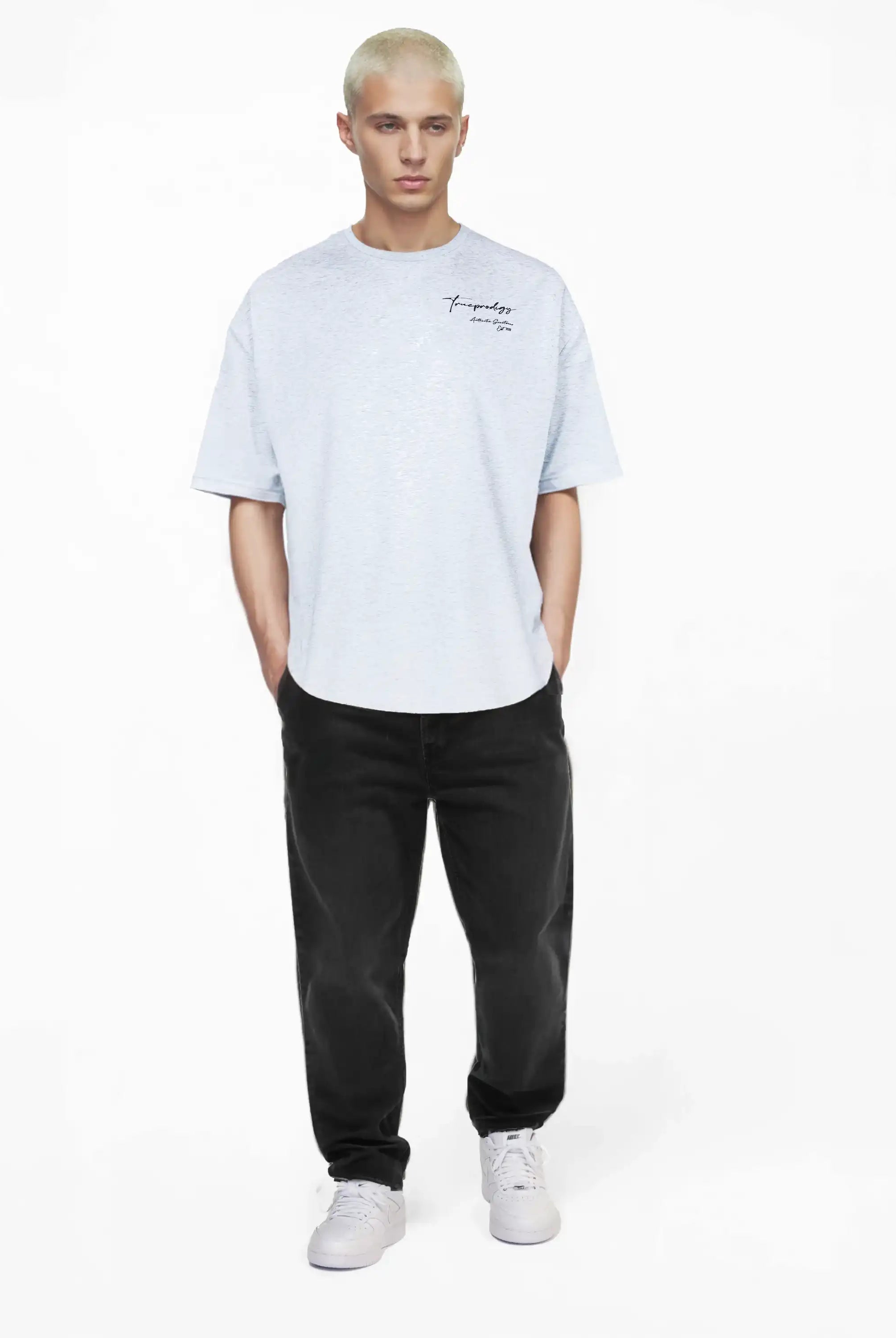 Eine Person mit kurzen gebleichten Haaren trägt das trueprodigy Marin Oversize-T-Shirt mit edlem Schriftzug auf der Brust in Hellgrau, gepaart mit schwarzen Jeans und weißen Turnschuhen, vor einem schlichten weißen Hintergrund.