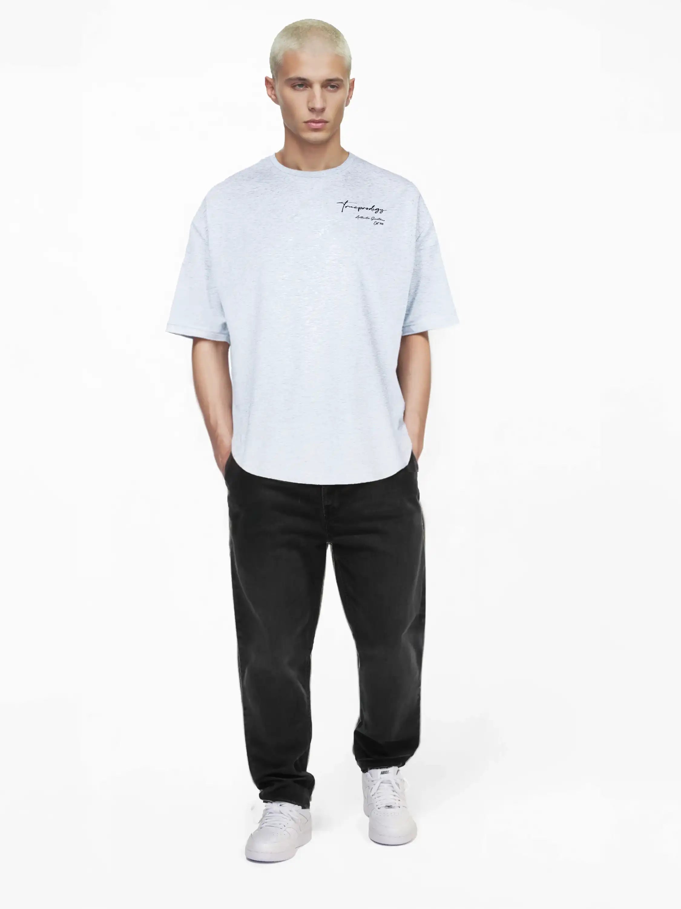 Eine Person mit kurzen gebleichten Haaren trägt das trueprodigy Marin Oversize-T-Shirt mit edlem Schriftzug auf der Brust in Hellgrau, gepaart mit schwarzen Jeans und weißen Turnschuhen, vor einem schlichten weißen Hintergrund.