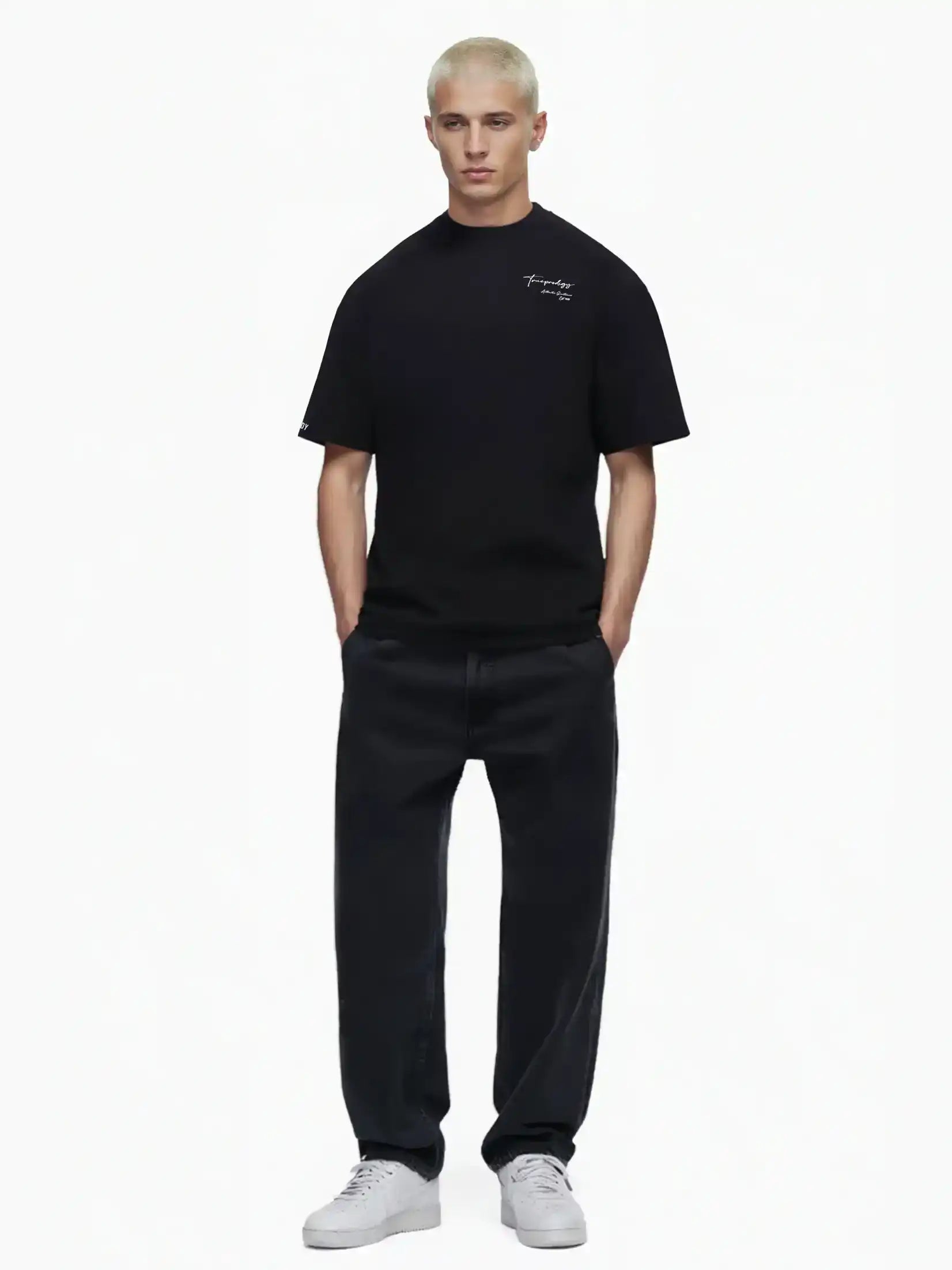 Eine Person mit kurzen blonden Haaren trägt das trueprodigy Marin Oversize-T-Shirt mit edlem Schriftzug auf der Brust, gepaart mit weiten schwarzen Jeans und weißen Turnschuhen, vor einem schlichten weißen Hintergrund.