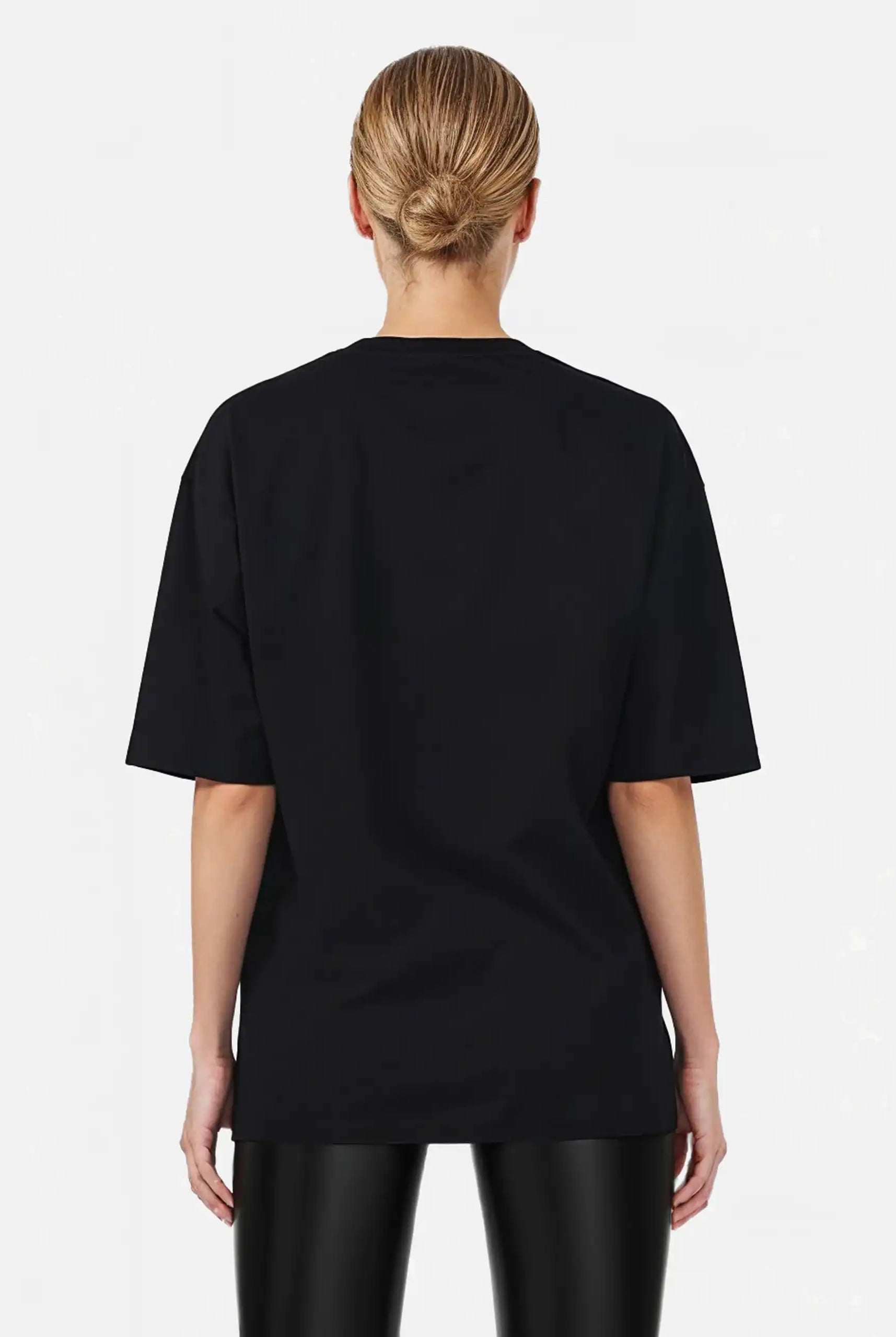 Eine Person mit blonden Haaren in einem tiefen Dutt ist von hinten zu sehen. Sie trägt das trueprodigy Marin F Oversize-T-Shirt mit feiner Logo-Schrift & TPDY-Print am Ärmel und eine schwarz glänzende Hose vor einem schlichten hellen Hintergrund.