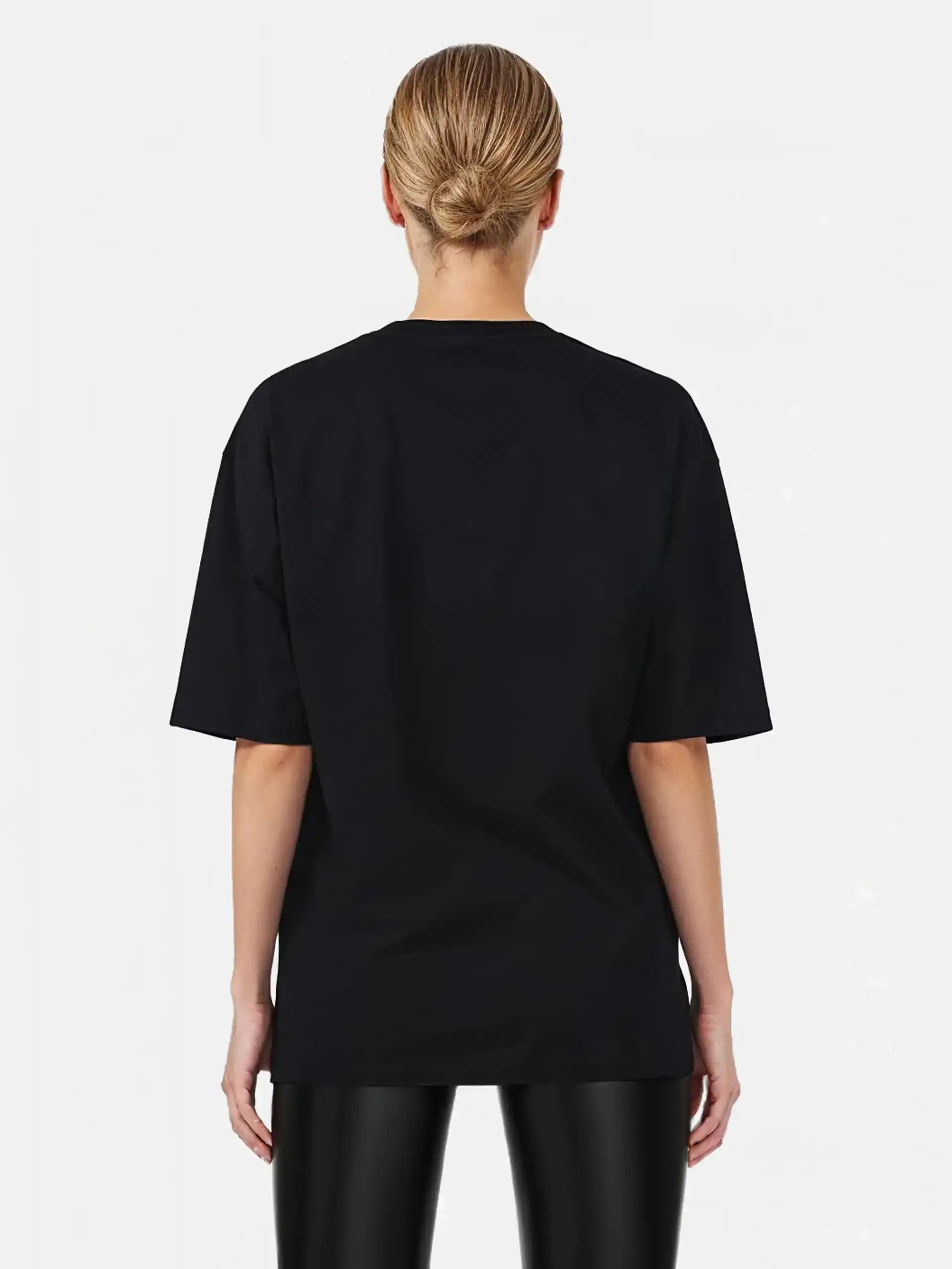 Eine Person mit blonden Haaren in einem tiefen Dutt ist von hinten zu sehen. Sie trägt das trueprodigy Marin F Oversize-T-Shirt mit feiner Logo-Schrift & TPDY-Print am Ärmel und eine schwarz glänzende Hose vor einem schlichten hellen Hintergrund.