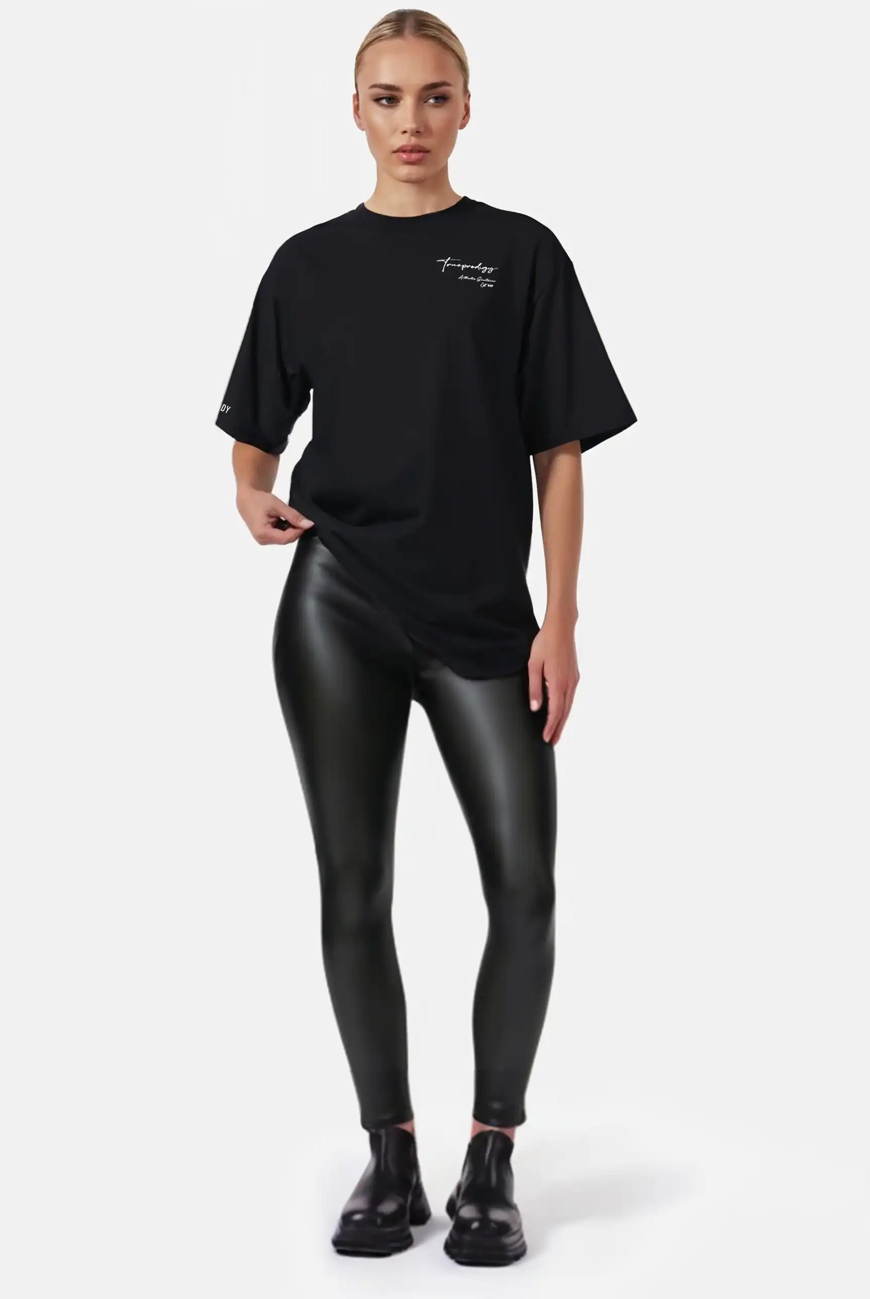 Eine Frau steht vor einem weißen Hintergrund und trägt das trueprodigy Marin F Oversize-T-Shirt mit feiner Logo-Schrift & TPDY-Print am Ärmel mit schwarz glänzenden Leggings und Schuhen. Ihr Haar ist zurückgesteckt und ihr Gesichtsausdruck ist neutral.