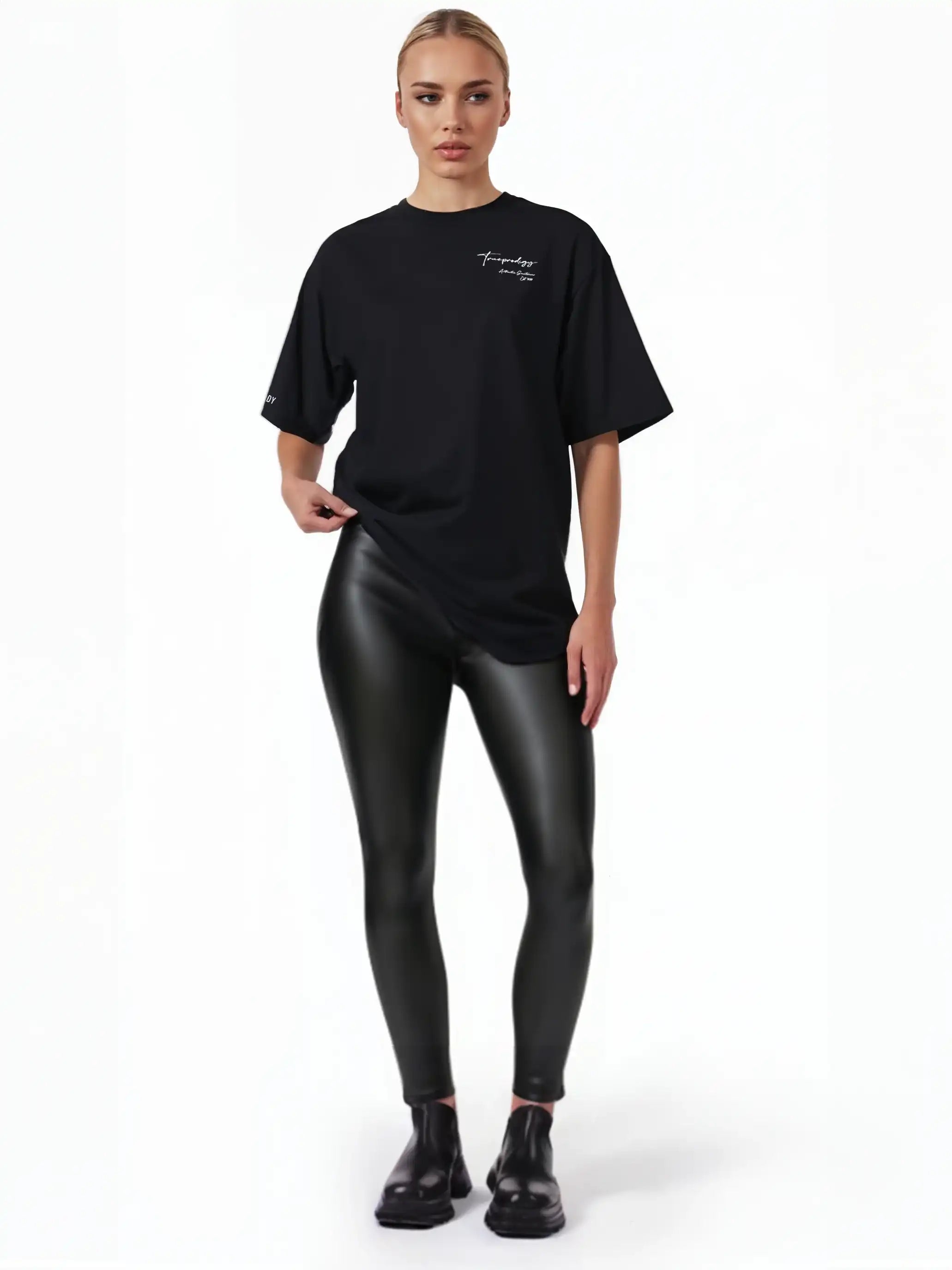 Eine Person steht vor einem weißen Hintergrund und trägt ein schwarzes trueprodigy Marin F Oversize-T-Shirt mit feiner Logo-Schrift & TPDY-Print am Ärmel, glänzende schwarze Leggings und schwarze Schuhe. Ihre hellen Haare sind zurückgesteckt und sie blicken neutral geradeaus.