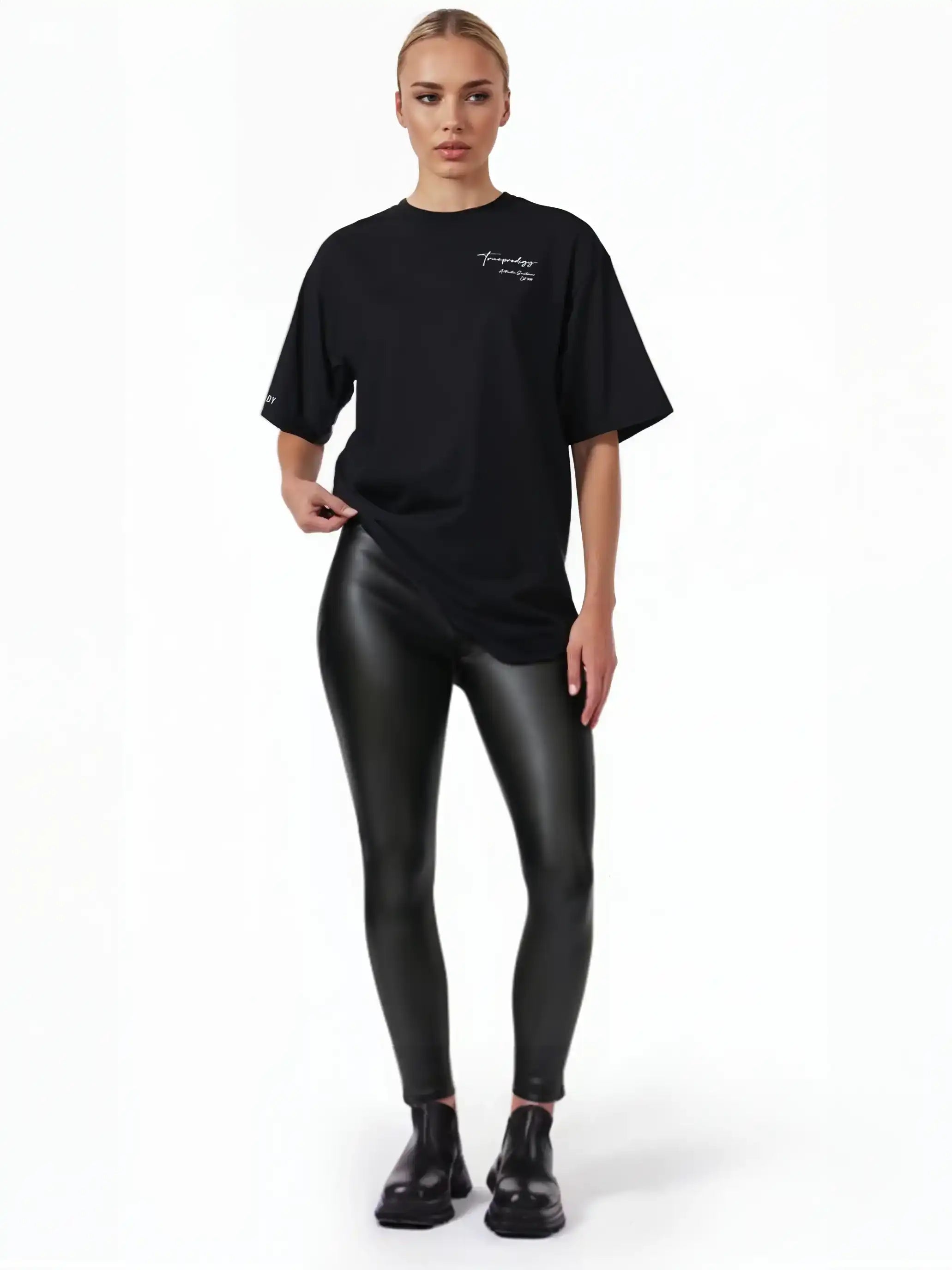 Eine Person steht vor einem weißen Hintergrund und trägt ein schwarzes trueprodigy Marin F Oversize-T-Shirt mit feiner Logo-Schrift & TPDY-Print am Ärmel, glänzende schwarze Leggings und schwarze Schuhe. Ihre hellen Haare sind zurückgesteckt und sie blicken neutral geradeaus.