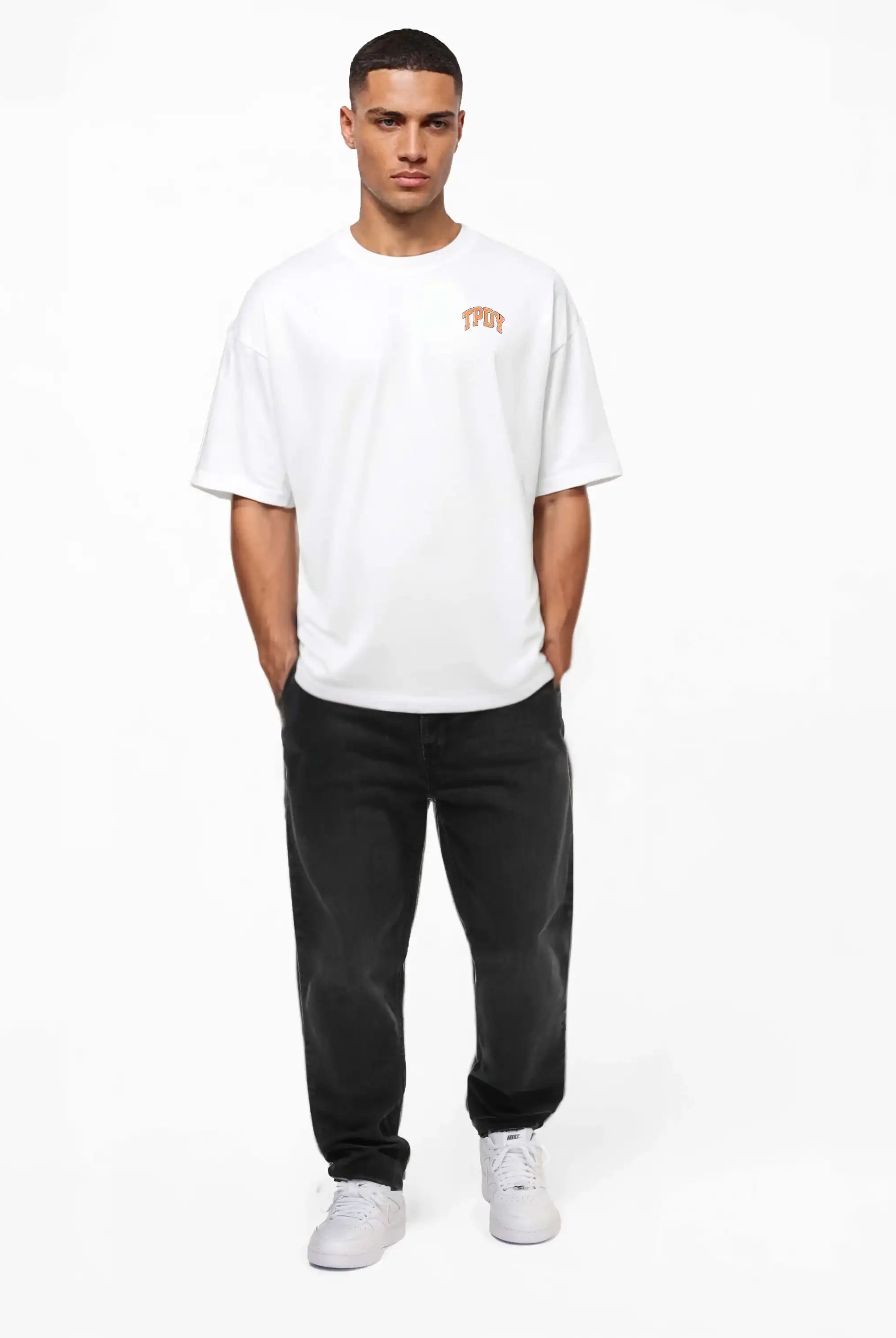 Ein Mann blickt nach vorne und trägt ein weißes trueprodigy Oversize-Shirt mit TPDY-Logo & Rückenprint "Stay Real", schwarze Jeans und weiße Turnschuhe. Er hat die Hände in den Taschen und steht vor einem schlichten weißen Hintergrund.