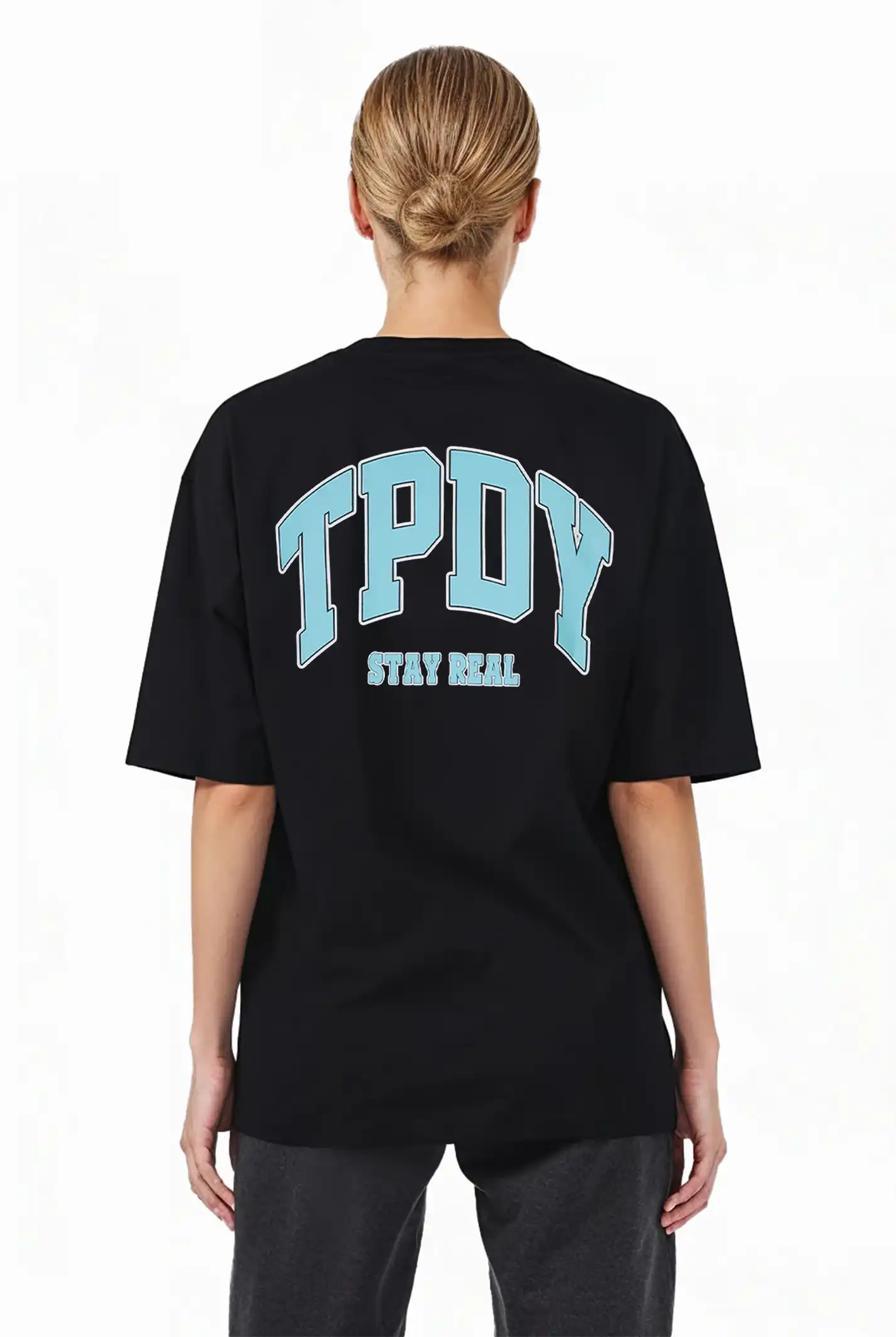 Eine Person mit blonden Haaren in einem tiefen Dutt, von der Kamera abgewandt, trägt das trueprodigy Arin F Oversize-T-Shirt mit TPDY-Print & "Stay Real" Rückenmotiv, aus dickerem Jersey Stoff, gepaart mit einer dunklen Hose.