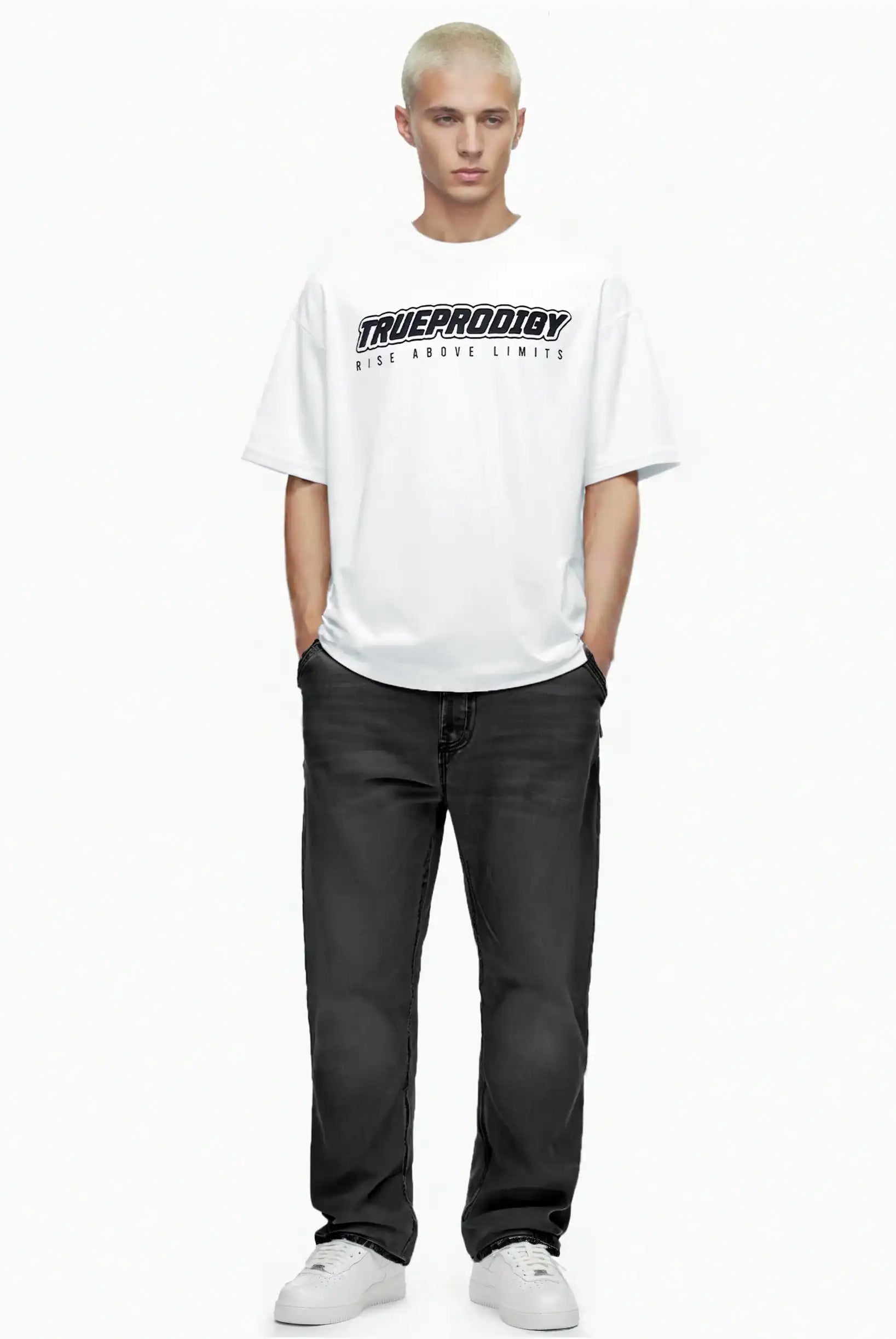 Eine Person mit kurzen blonden Haaren steht mit dem Blick nach vorne, trägt das trueprodigy Toni Oversize-Shirt mit 3D-Schaumprint & "Rise Above Limits", schwarze Jeans und weiße Turnschuhe. Ihre Hände stecken in den Taschen vor einem schlichten weißen Hintergrund.