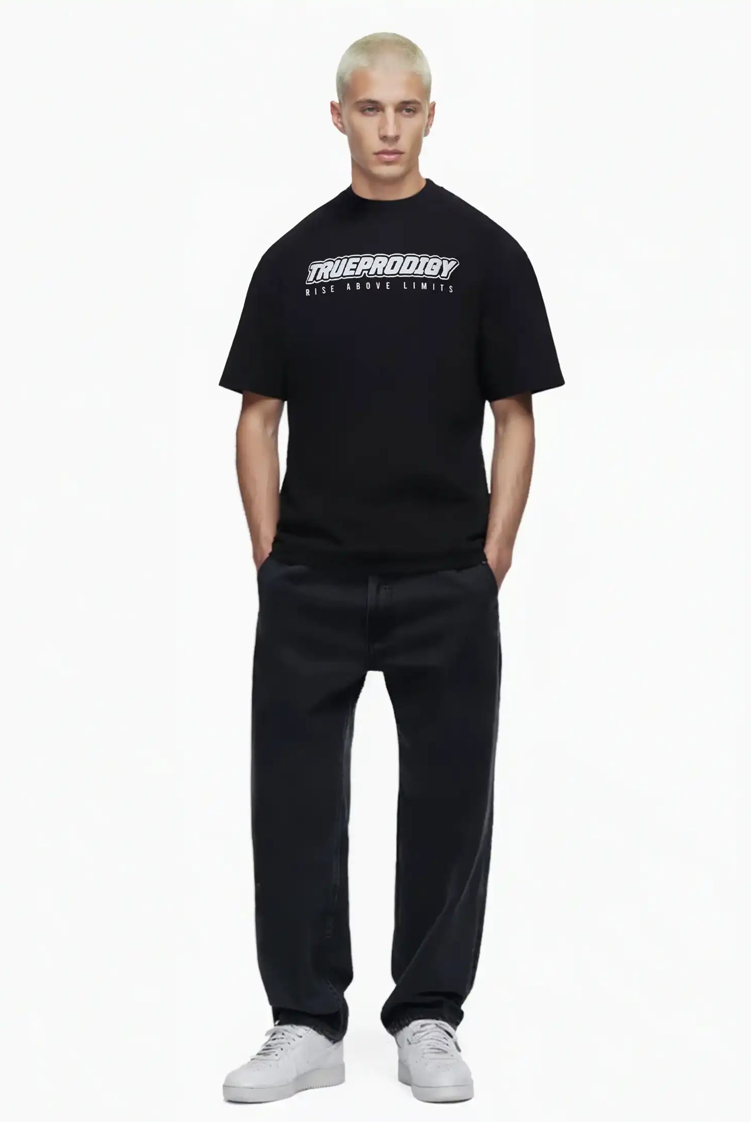 Eine Person mit kurzen, gebleichten blonden Haaren trägt das trueprodigy Toni Oversize-Shirt mit 3D-Schaumprint & "Rise Above Limits", lockere schwarze Jeans und weiße Turnschuhe, stehend vor einem schlichten weißen Hintergrund.
