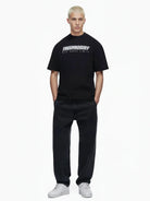 Eine Person mit kurzen, gebleichten blonden Haaren trägt das trueprodigy Toni Oversize-Shirt mit 3D-Schaumprint & "Rise Above Limits", lockere schwarze Jeans und weiße Turnschuhe, stehend vor einem schlichten weißen Hintergrund.