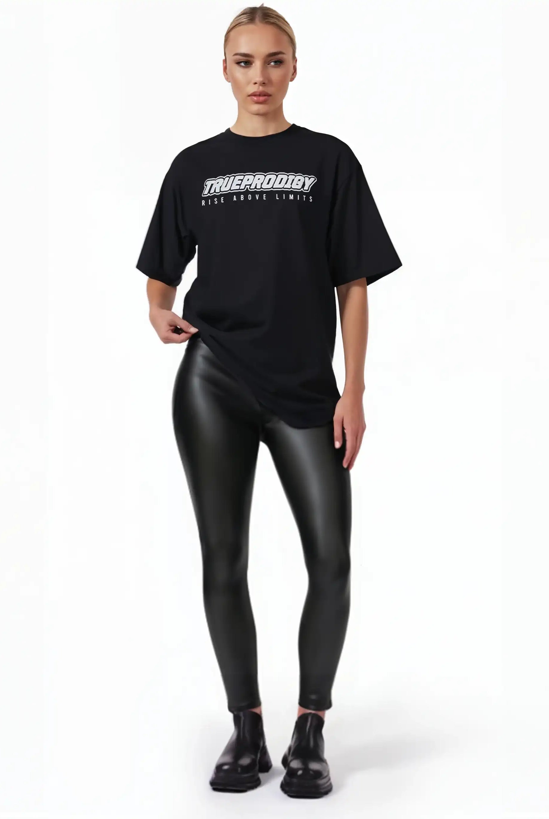 Eine Person steht vor einem schlichten weißen Hintergrund und trägt das trueprodigy Toni F Oversize-T-Shirt mit 3D-Schriftzug "TRUEPRODIGY" & "Rise Above Limits", gepaart mit schwarzen Kunstlederleggings und schwarzen Schuhen. Die Haare sind hochgesteckt und eine Hand hält den Hemdsaum fest.