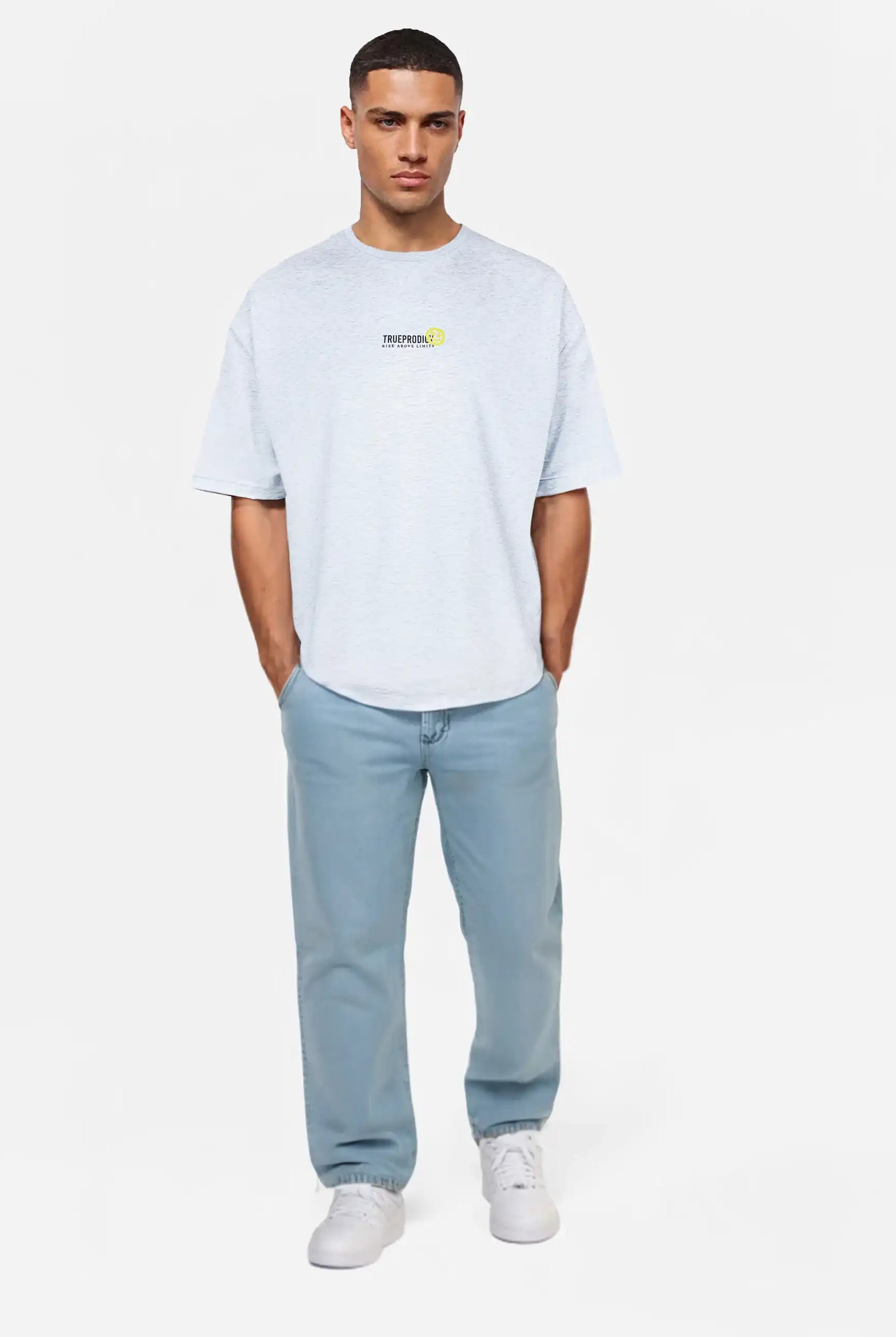 Ein Mann blickt nach vorne in einem Trueprodigy Oversize-Herren-T-Shirt mit Logo-Smile-Grafik auf der Brust, hellblauen Jeans und weißen Sneakers. Seine Hände stecken in den Taschen vor einem schlichten weißen Hintergrund.