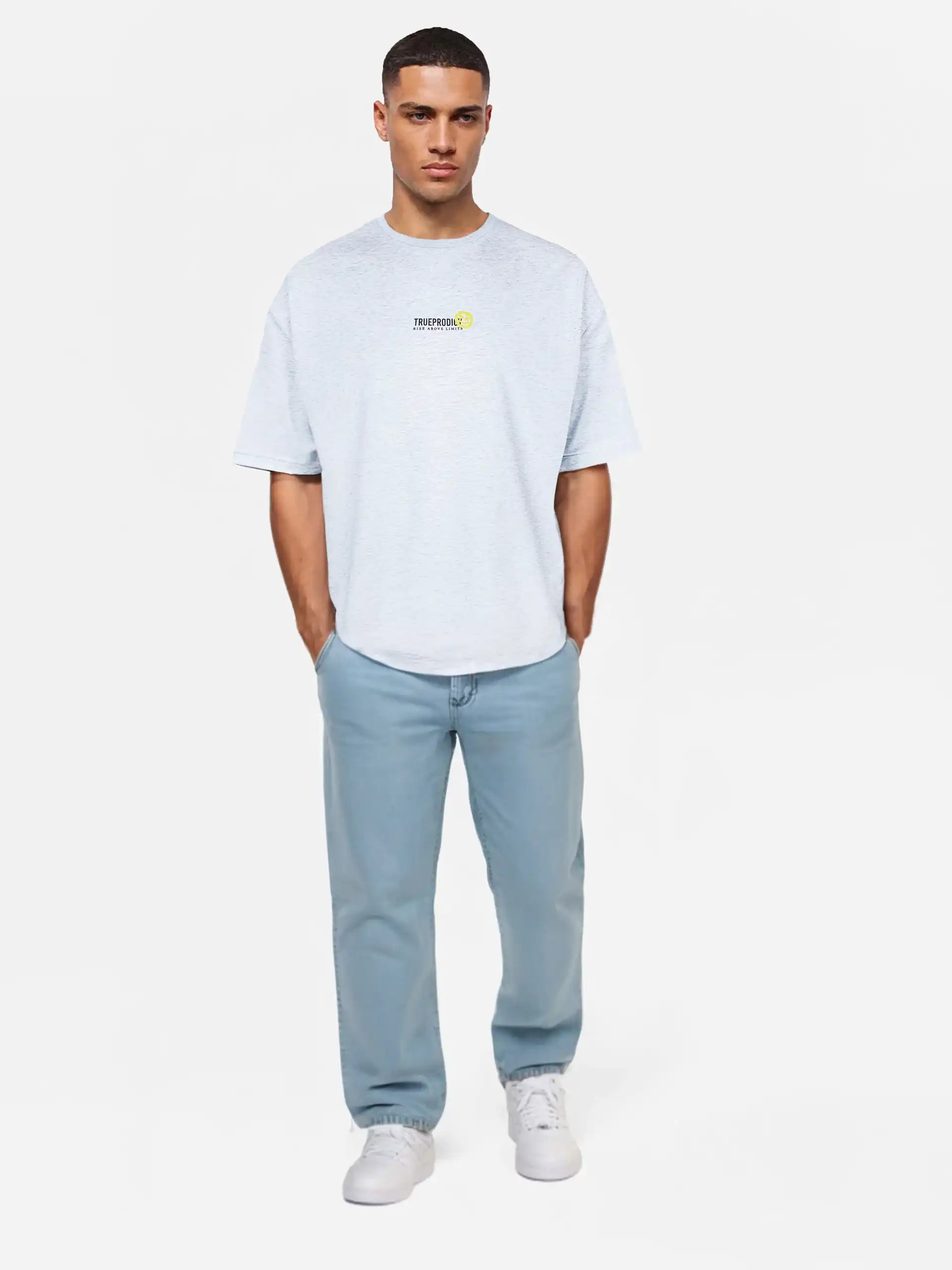 Ein Mann blickt nach vorne in einem Trueprodigy Oversize-Herren-T-Shirt mit Logo-Smile-Grafik auf der Brust, hellblauen Jeans und weißen Sneakers. Seine Hände stecken in den Taschen vor einem schlichten weißen Hintergrund.