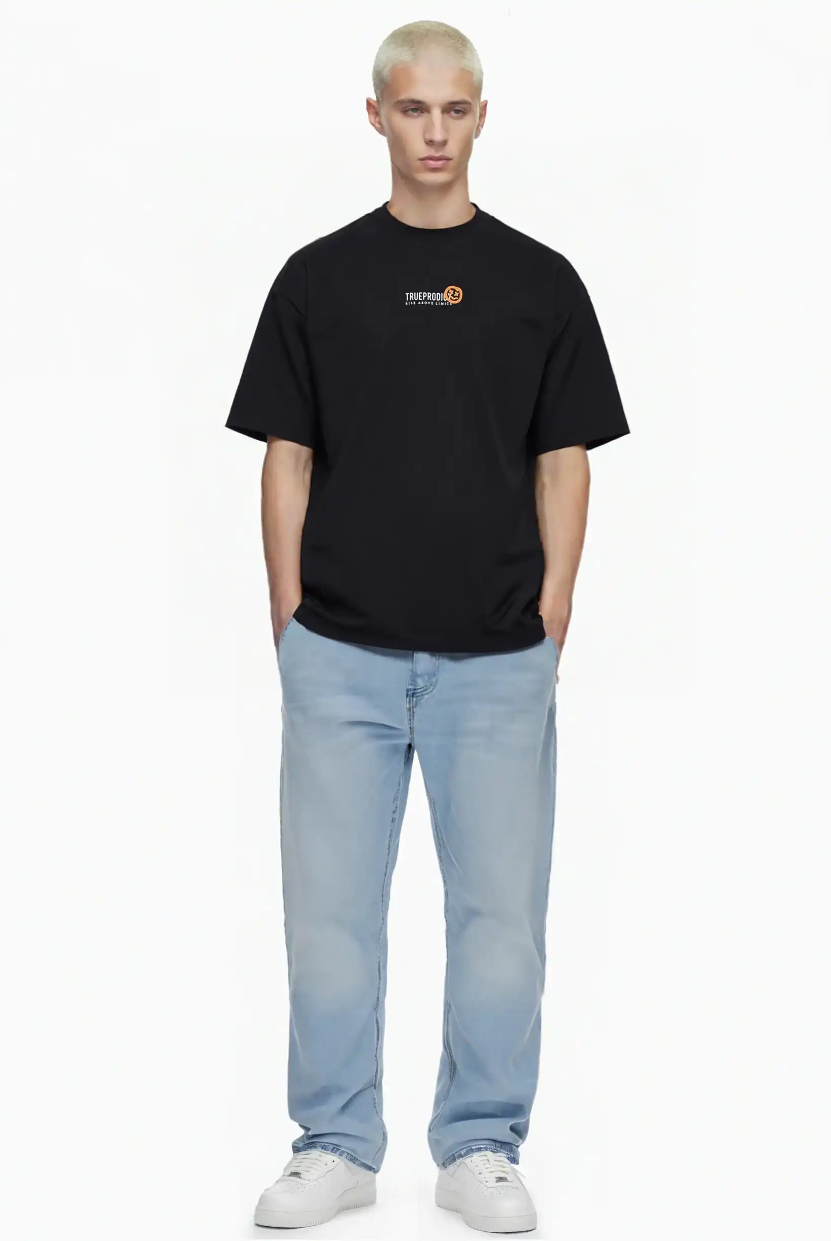 Eine Person mit kurzen blonden Haaren steht vor einem weißen Hintergrund und trägt das trueprodigy Juniper Oversize-Herren-T-Shirt mit Logo-Smile-Grafik auf der Brust, hellblaue Jeans und weiße Turnschuhe. Die Hände stecken in den Taschen.