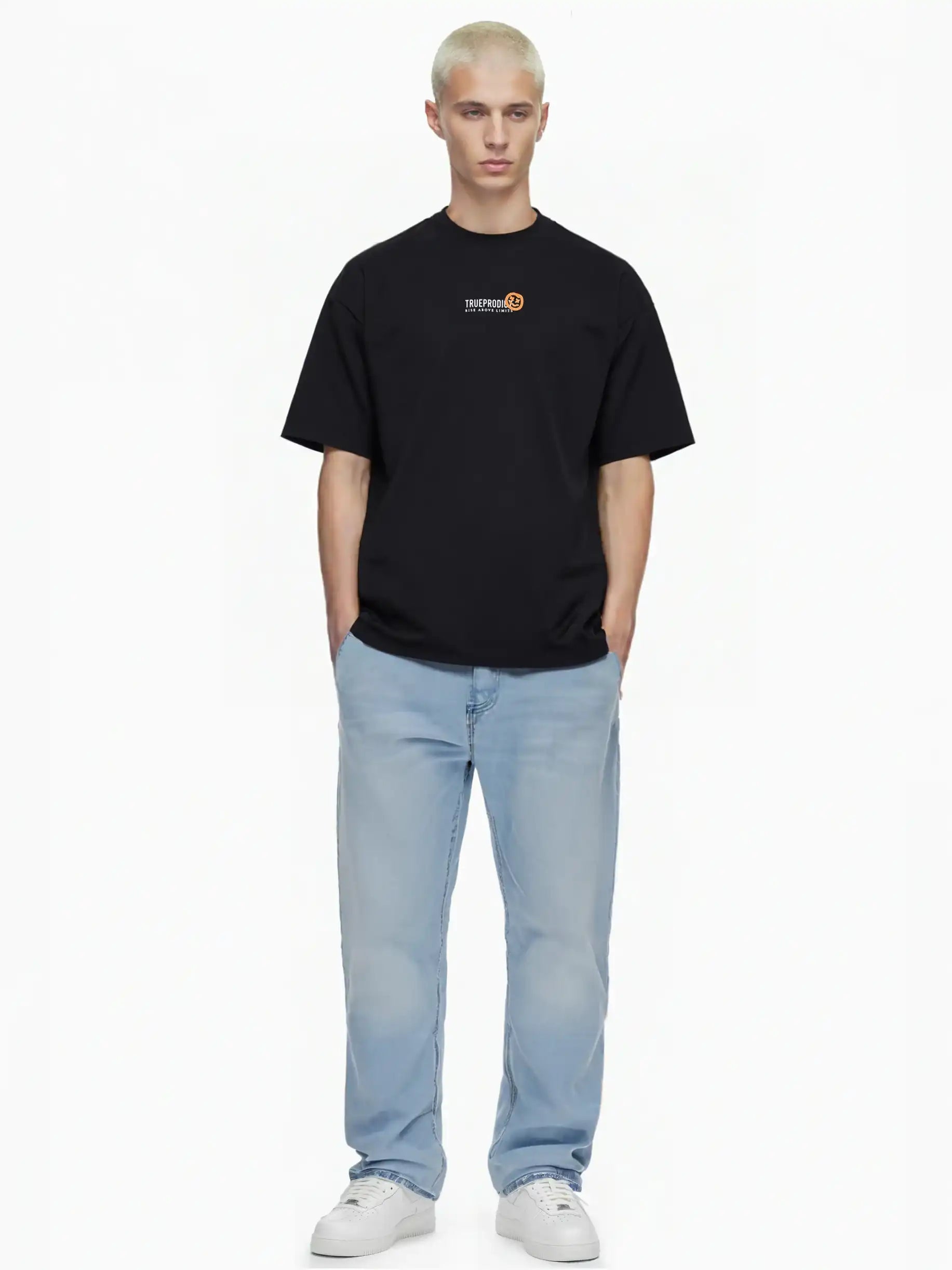 Eine Person mit kurzen blonden Haaren steht vor einem weißen Hintergrund und trägt das trueprodigy Juniper Oversize-Herren-T-Shirt mit Logo-Smile-Grafik auf der Brust, hellblaue Jeans und weiße Turnschuhe. Die Hände stecken in den Taschen.