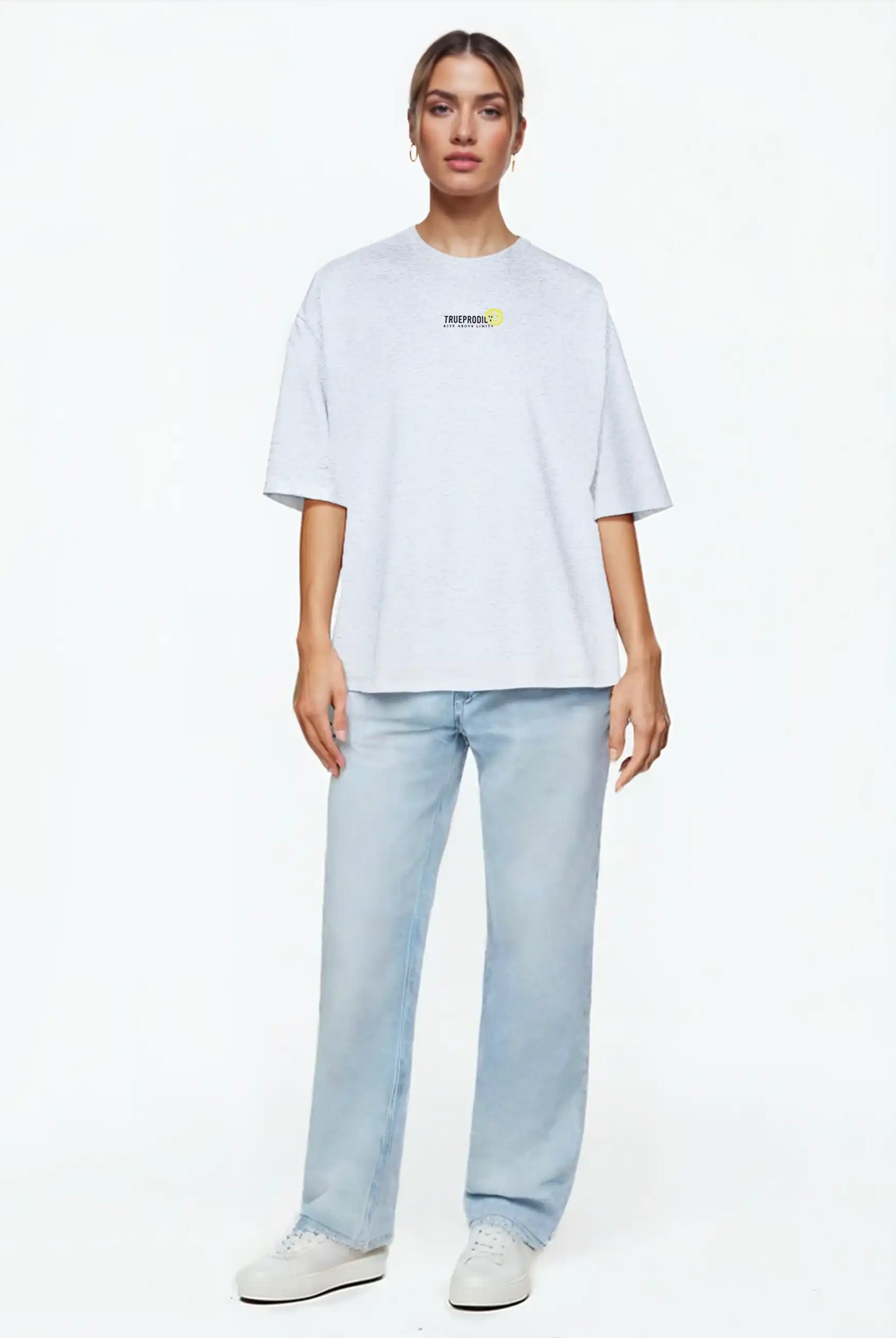 Eine Person steht vor einem weißen Hintergrund und trägt das trueprodigy Juniper F Oversize-Damen-T-Shirt mit Logo-Smile-Grafik auf der Vorderseite, gepaart mit hellblauen Straight-Leg-Jeans und weißen Sneakers.