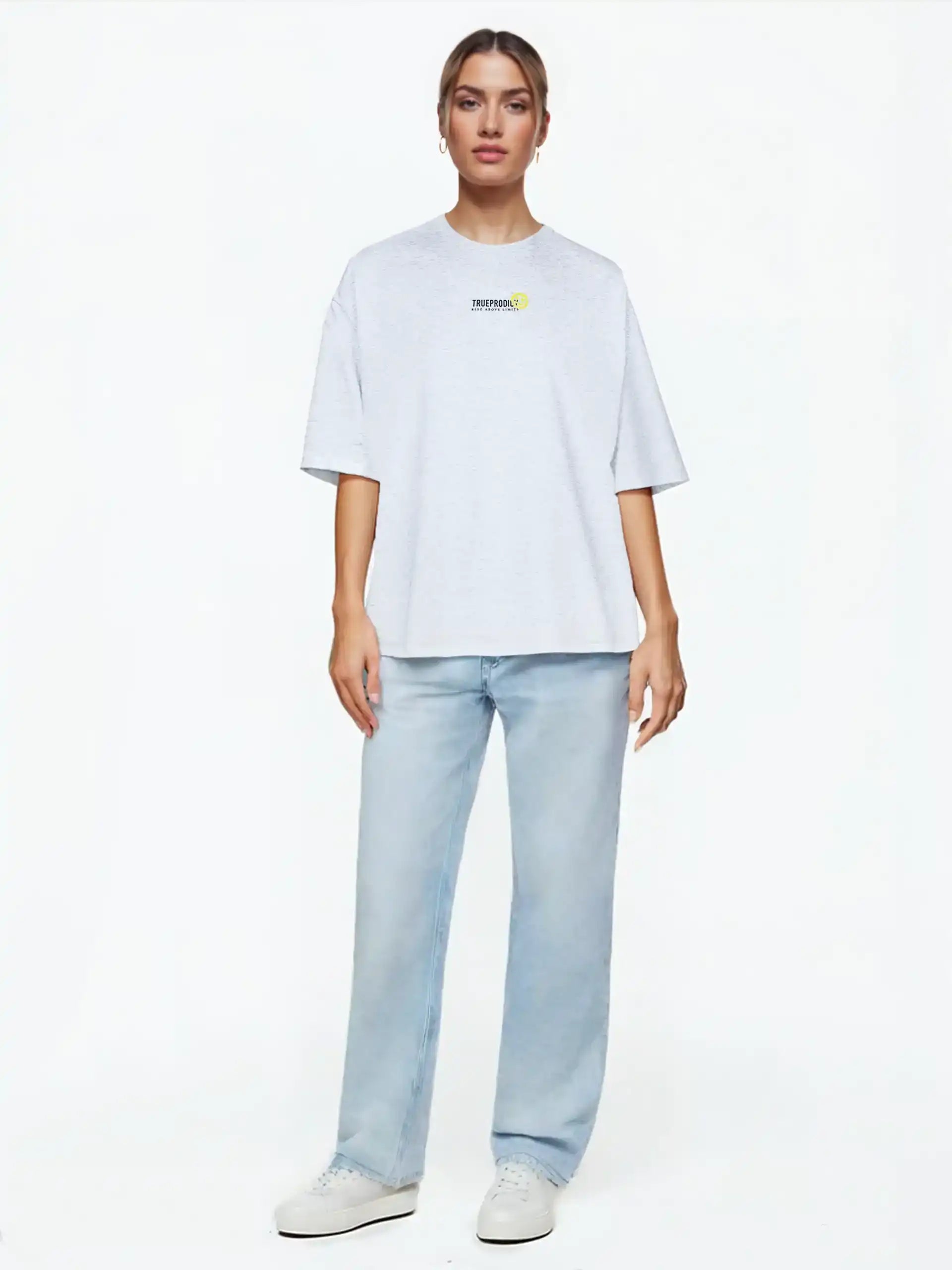 Eine Person steht vor einem weißen Hintergrund und trägt das trueprodigy Juniper F Oversize-Damen-T-Shirt mit Logo-Smile-Grafik auf der Vorderseite, gepaart mit hellblauen Straight-Leg-Jeans und weißen Sneakers.