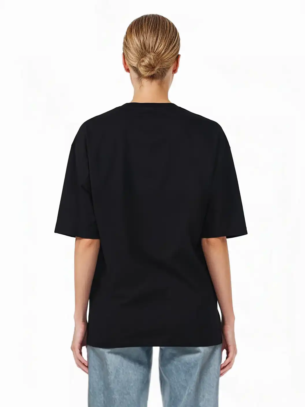 Eine Person mit blondem Haar in einem tiefen Dutt, die von der Kamera wegschaut, trägt ein schwarzes trueprodigy Juniper F Oversize-Damen-T-Shirt mit Logo-Smile-Grafik auf der Vorderseite und hellblaue Jeans vor einem weißen Hintergrund.