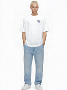 Eine Person mit kurzen blonden Haaren steht vor einem weißen Hintergrund und trägt das trueprodigy Hajin Oversize-T-Shirt mit Spray-Smiley & Rückenprint "Happy Mind", hellblaue Jeans und weiße Sneakers, die Hände lässig in den Taschen.