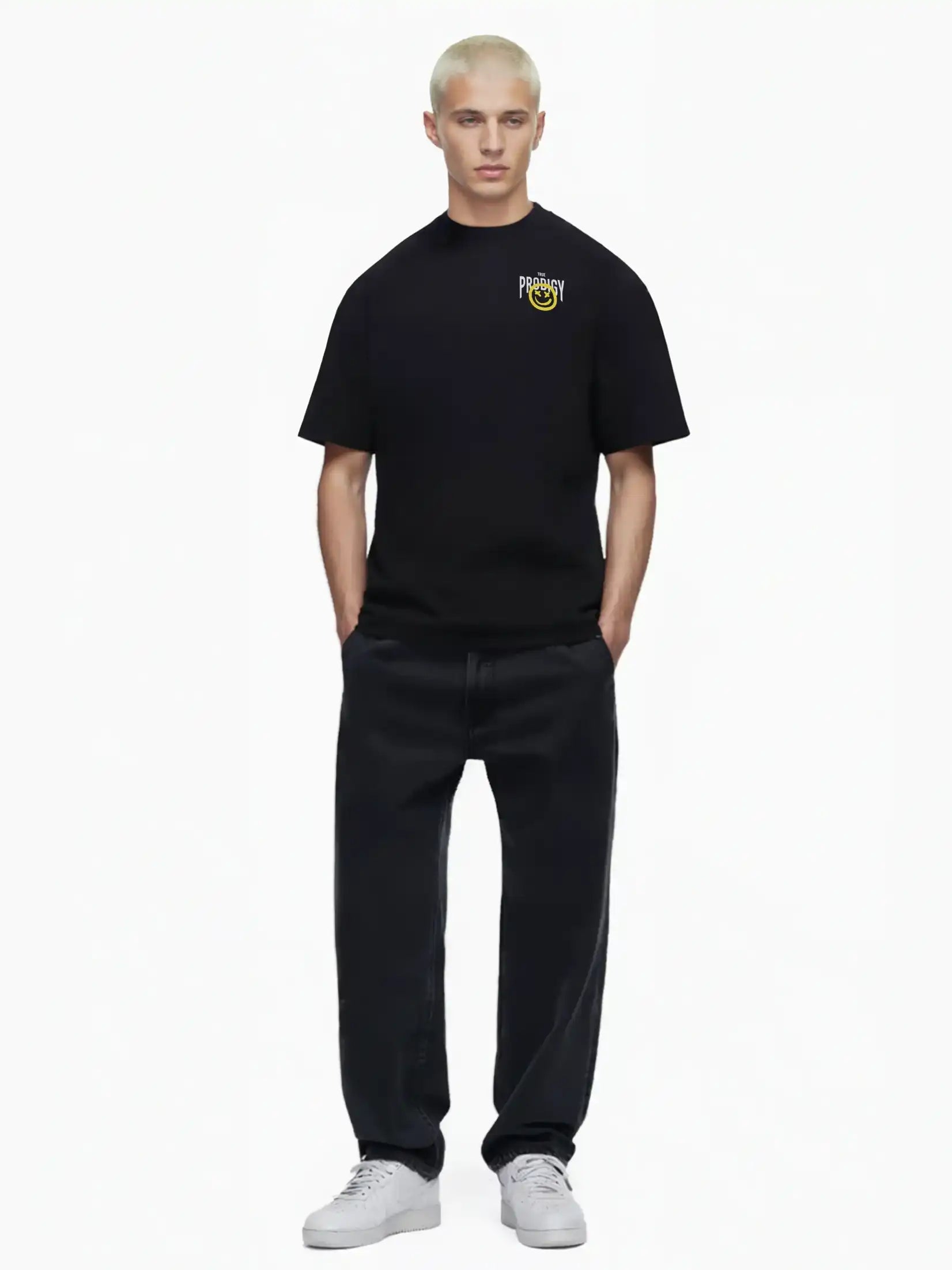 Eine Person mit kurzen blonden Haaren blickt nach vorne und trägt das trueprodigy Hajin Oversize-T-Shirt mit Spray-Smiley & Rückenprint "Happy Mind", gepaart mit schwarzen Jeans und weißen Turnschuhen, vor einem schlichten weißen Hintergrund.