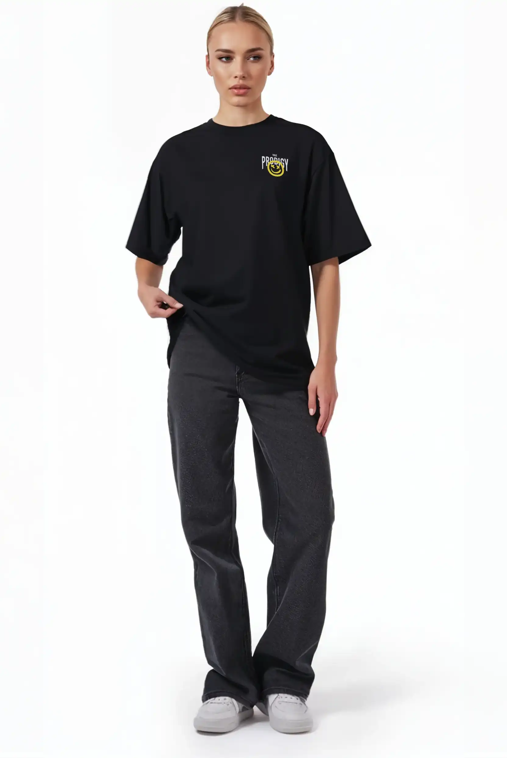 Eine Person steht vor einem weißen Hintergrund und trägt das trueprodigy Hajin F Oversize-T-Shirt mit Spray-Smiley & Happy-Mind-Print, gepaart mit dunklen Jeans und weißen Sneakers. Eine Hand hält die Seite des Shirts; der Gesichtsausdruck ist neutral.
