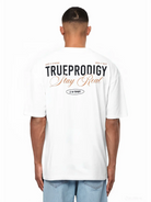 Eine Person schaut weg und trägt das trueprodigy Lou Oversize-T-Shirt aus schwerem Jerseystoff mit auffälligen "Stay Real"- und Schriftzug-Prints auf Vorder- und Rückseite, gepaart mit hellen Denim-Shorts.