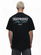 Eine Person mit kurzen blonden Haaren trägt von hinten das trueprodigy Lou Oversize-T-Shirt mit Schriftzug & "Stay Real" Front- & Rückenprint, mit weißem und blauem Druck auf dem oberen Rücken.