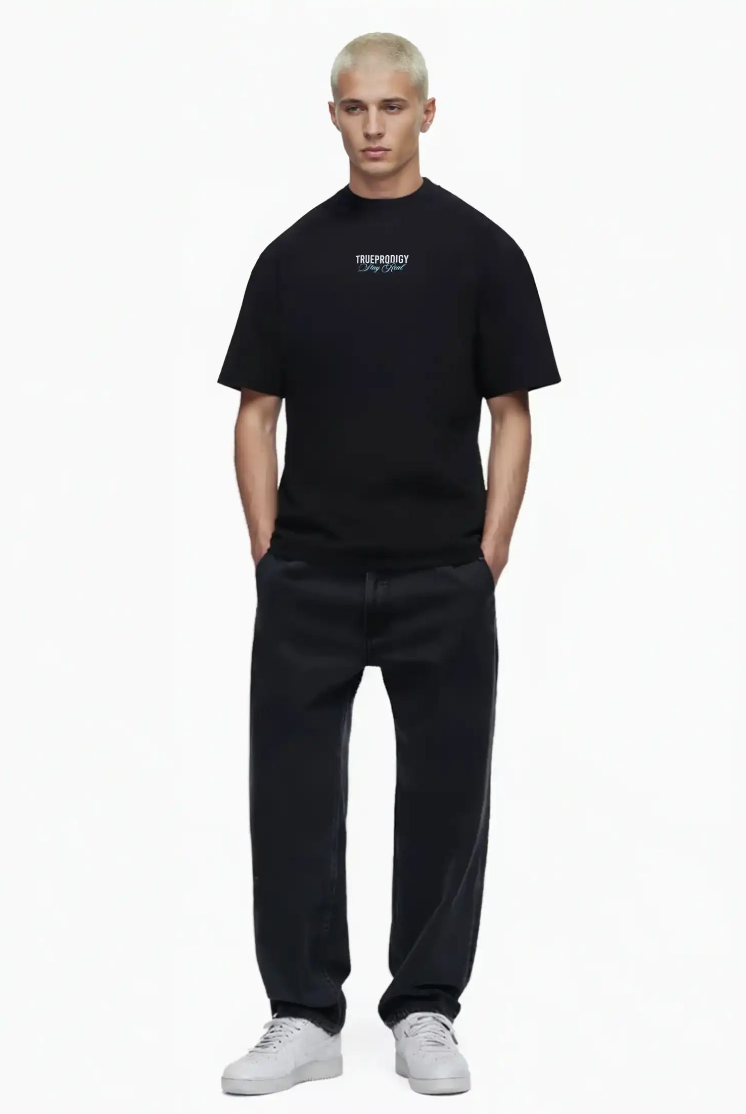 Eine Person mit kurzen blonden Haaren steht vor einem weißen Hintergrund und trägt das trueprodigy Lou Oversize-T-Shirt mit Schriftzug & "Stay Real" Front- & Rückenprint, weite schwarze Jeans und weiße Turnschuhe. Die Hände stecken in den Taschen.
