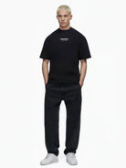 Eine Person mit kurzen blonden Haaren steht vor einem weißen Hintergrund und trägt das trueprodigy Lou Oversize-T-Shirt mit Schriftzug & "Stay Real" Front- & Rückenprint, weite schwarze Jeans und weiße Turnschuhe. Die Hände stecken in den Taschen.