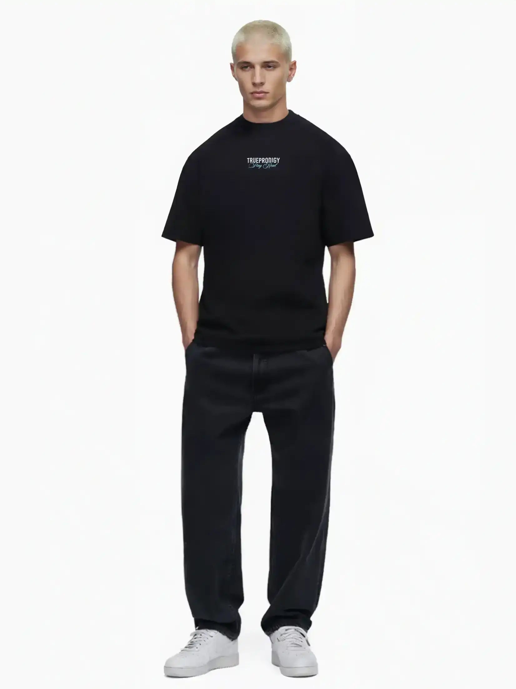 Eine Person mit kurzen blonden Haaren steht vor einem weißen Hintergrund und trägt das trueprodigy Lou Oversize-T-Shirt mit Schriftzug & "Stay Real" Front- & Rückenprint, weite schwarze Jeans und weiße Turnschuhe. Die Hände stecken in den Taschen.