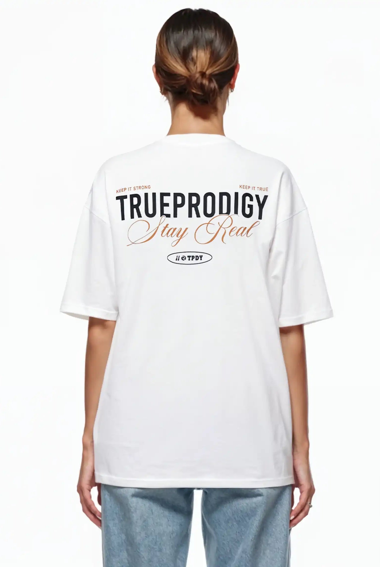 Eine Person blickt von der Kamera weg und trägt ein weißes trueprodigy Lou F Oversize-T-Shirt mit dem Schriftzug "Stay Real" auf Vorder- und Rückseite, gepaart mit einer hellblauen Jeans. Die geräumige Passform hebt sich von dem schlichten weißen Hintergrund ab.