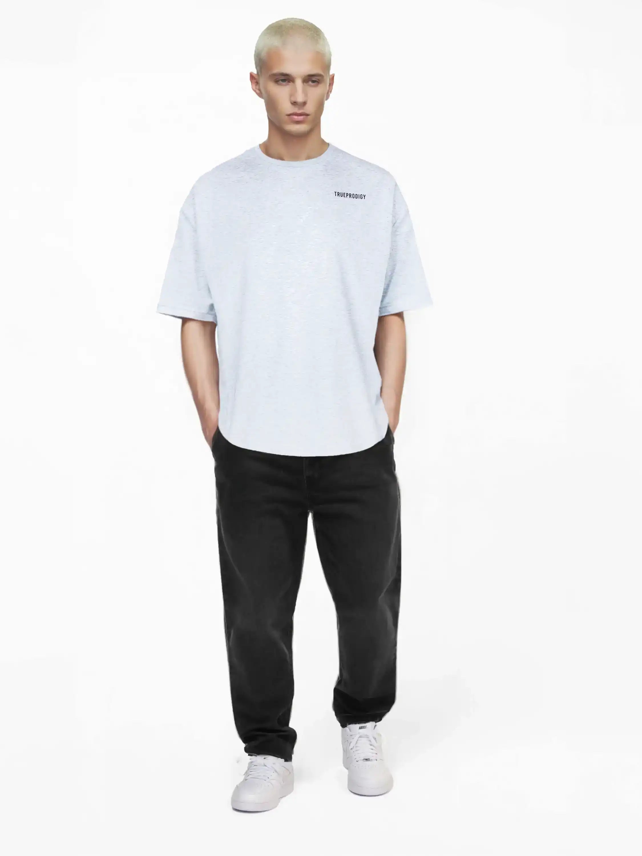 Eine Person mit kurzen, hellblonden Haaren trägt das trueprodigy Lior Oversize-T-Shirt mit Brustlogo und Rückenprint "never back down" in Hellgrau, gepaart mit schwarzen Jeans und weißen Turnschuhen, und steht mit dem Gesicht nach vorne vor einem schlichten weißen Hintergrund.