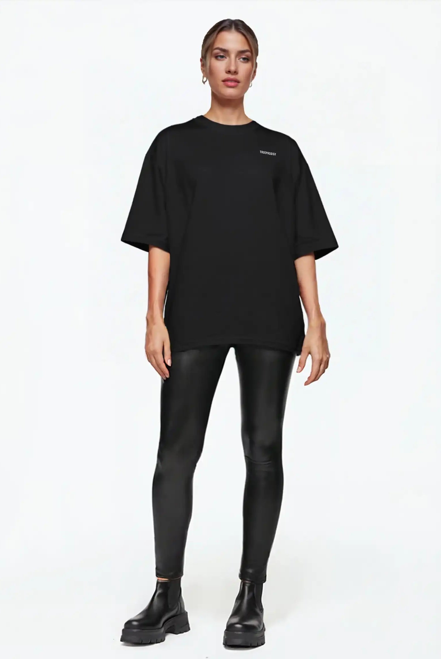Eine Person steht mit dem Gesicht nach vorne und trägt ein trueprodigy Lior F Oversize-Damen-T-Shirt mit Rückenstatement "never back down", schwarz glänzende Leggings und Ankle Boots vor einem schlichten weißen Hintergrund.