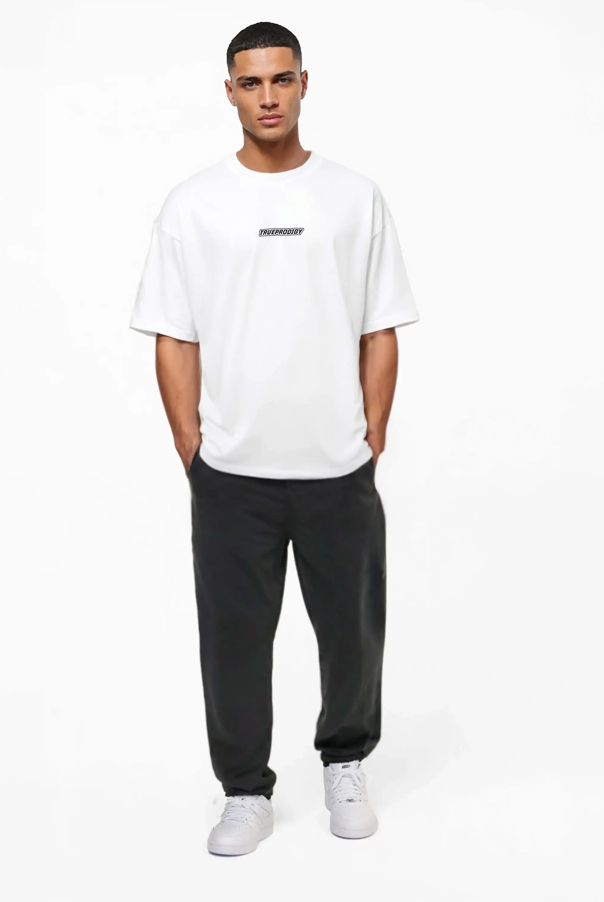 Eine Person steht vor einem schlichten weißen Hintergrund und trägt das Eden Oversize-Shirt mit 3D-Logo-Schaumprint auf Brust & Rücken von trueprodigy, gepaart mit einer schwarzen Hose und weißen Sneakers, die Hände in den Taschen.