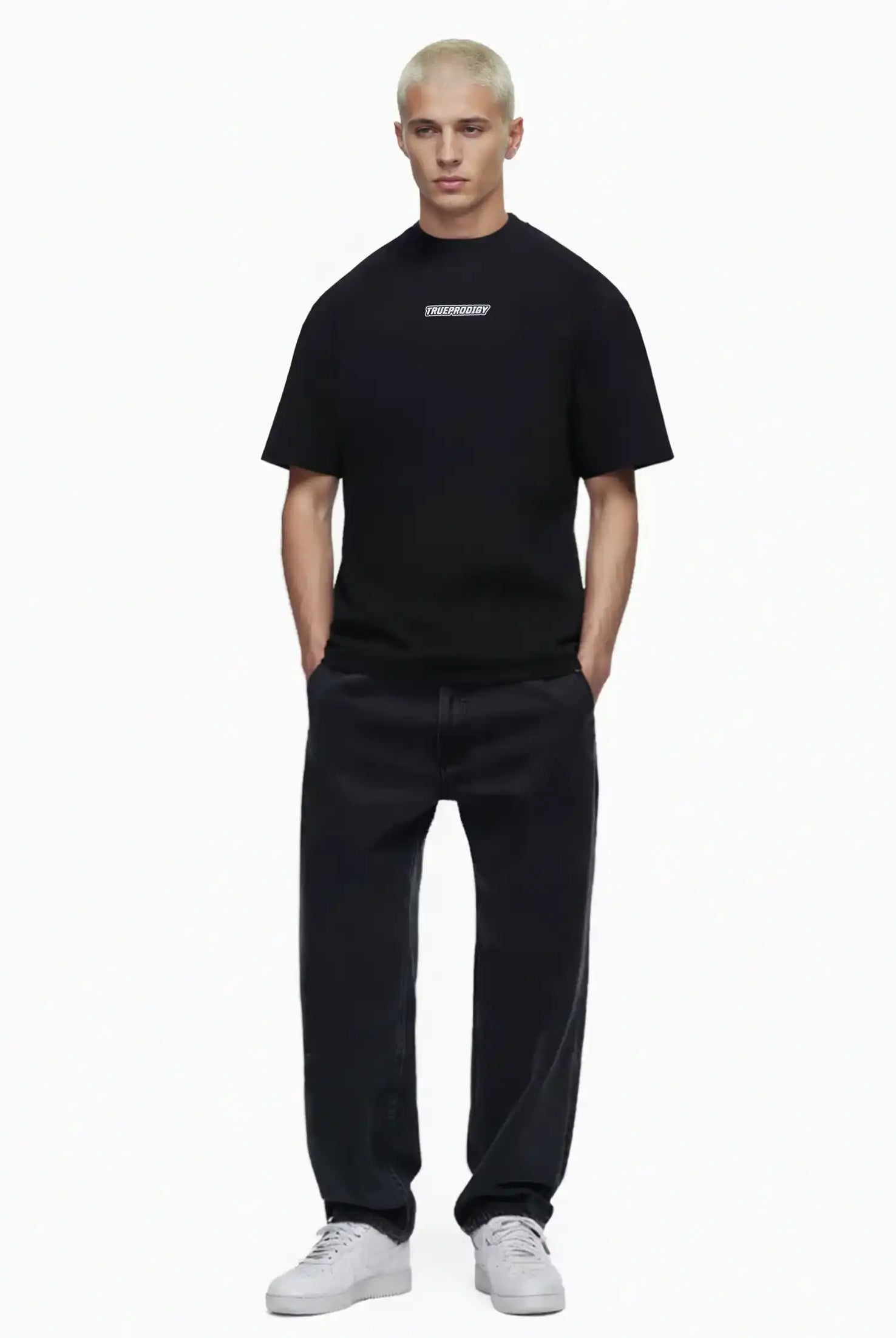 Eine Person mit kurzen blonden Haaren blickt nach vorne und trägt ein schwarzes trueprodigy Eden Oversize-Shirt mit 3D-Logo-Schaumprint auf Brust & Rücken, weite schwarze Jeans und weiße Turnschuhe vor einem schlichten weißen Hintergrund.