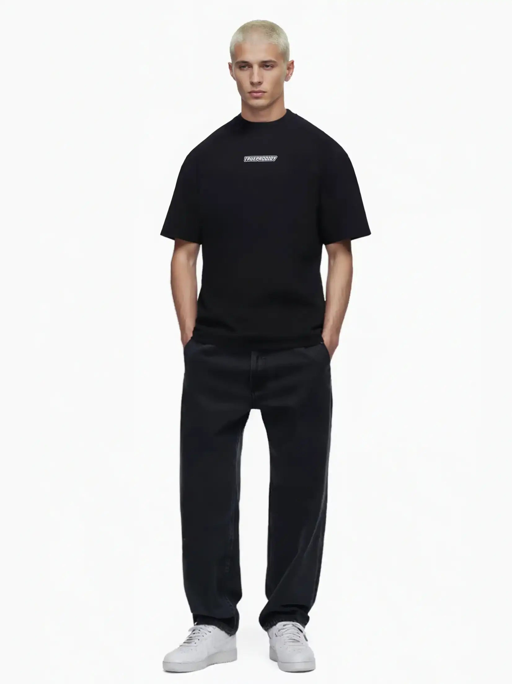 Eine Person mit kurzen blonden Haaren blickt nach vorne und trägt ein schwarzes trueprodigy Eden Oversize-Shirt mit 3D-Logo-Schaumprint auf Brust & Rücken, weite schwarze Jeans und weiße Turnschuhe vor einem schlichten weißen Hintergrund.
