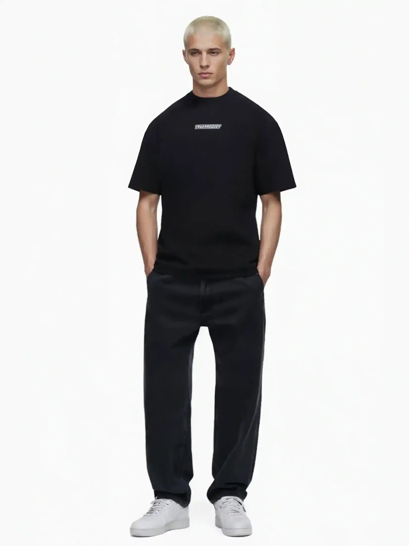 Eine Person mit kurzen blonden Haaren blickt nach vorne und trägt ein schwarzes trueprodigy Eden Oversize-Shirt mit 3D-Logo-Schaumprint auf Brust & Rücken, weite schwarze Jeans und weiße Turnschuhe vor einem schlichten weißen Hintergrund.