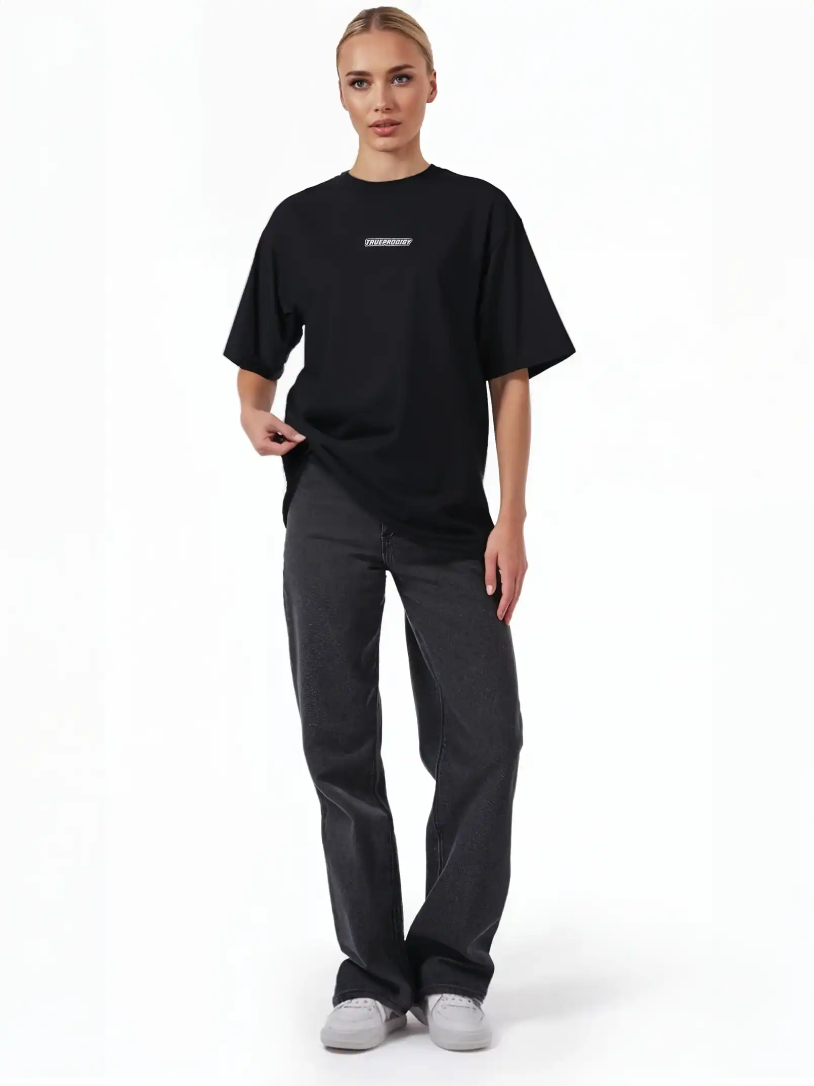Eine Person steht mit dem Blick nach vorne und trägt das schwarze trueprodigy Eden F Oversize-T-Shirt mit 3D-Logo-Schriftzug vorn & großem Rückenprint, gepaart mit dunklen Straight-Leg-Jeans und weißen Sneakers vor einem schlichten weißen Hintergrund.