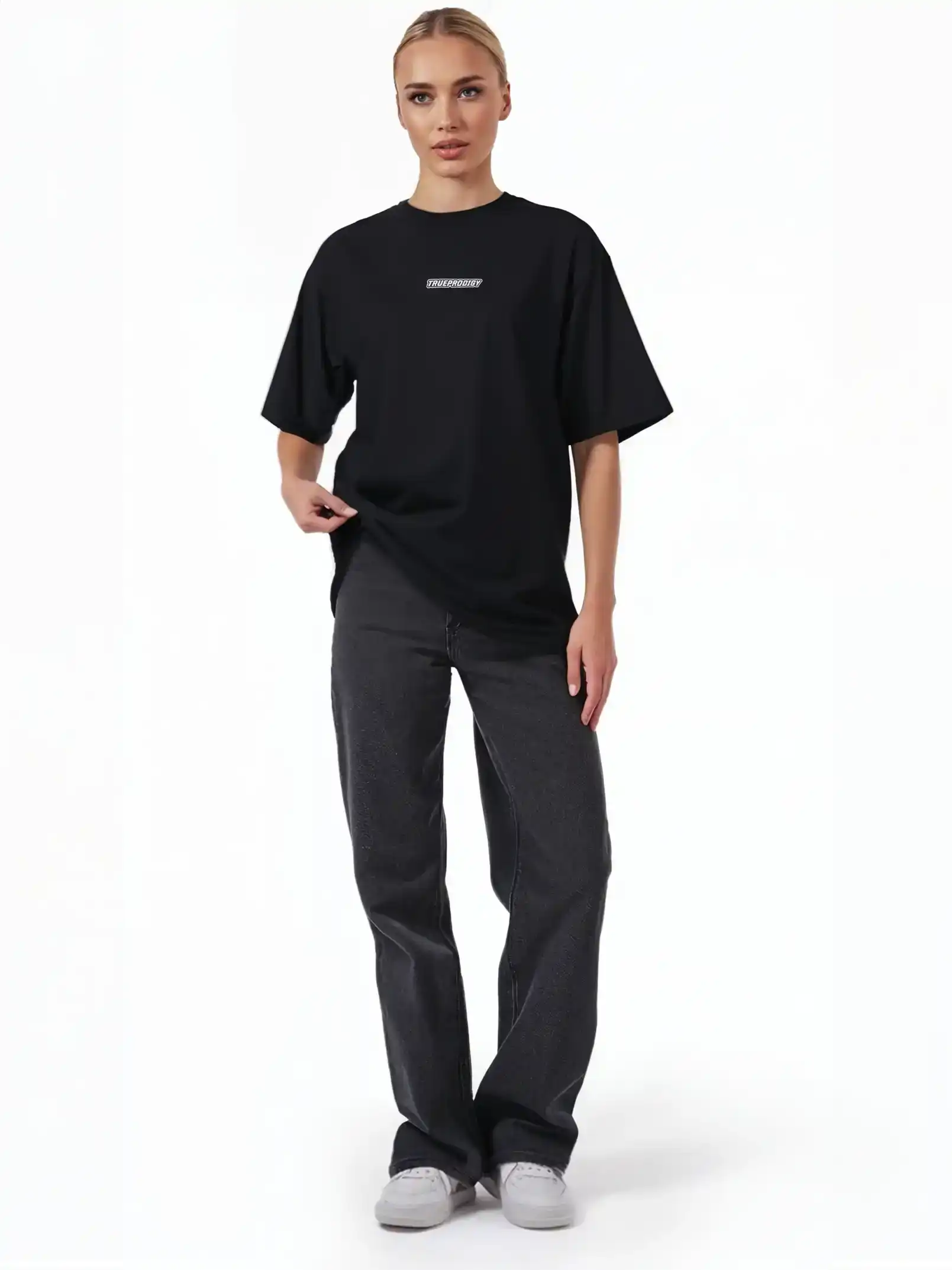 Eine Person steht mit dem Blick nach vorne und trägt das schwarze trueprodigy Eden F Oversize-T-Shirt mit 3D-Logo-Schriftzug vorn & großem Rückenprint, gepaart mit dunklen Straight-Leg-Jeans und weißen Sneakers vor einem schlichten weißen Hintergrund.