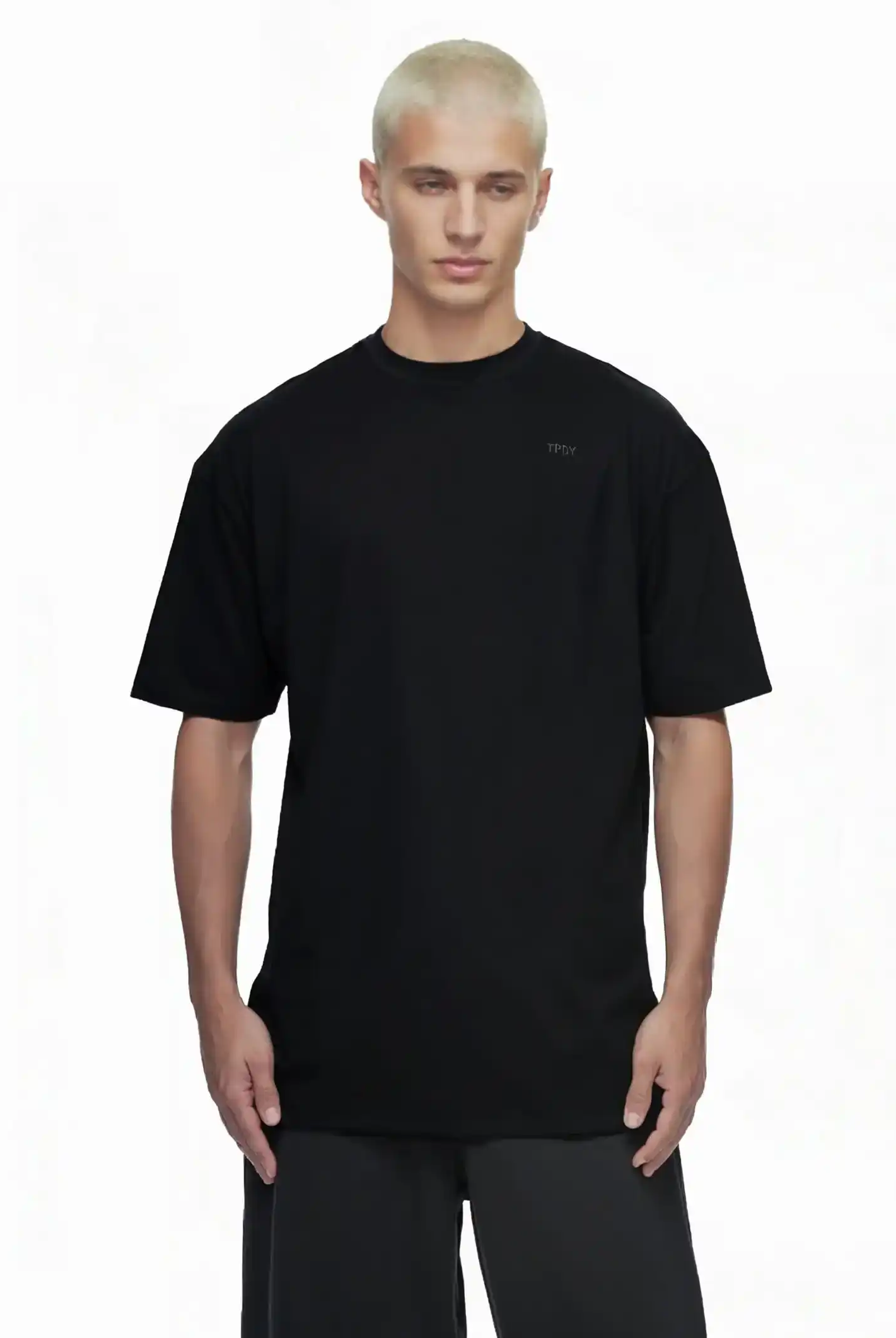 Eine Person mit kurzen, gebleichten blonden Haaren trägt das trueprodigy Haram Oversize-Herren-T-Shirt mit dezentem TPDY-Sticklogo in Schwarz und schwarzer Hose vor weißem Hintergrund. Das Shirt hat eine dezente Bruststickerei.